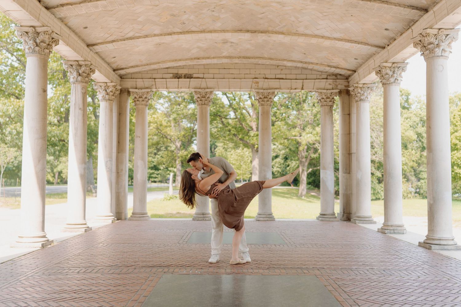 Couple in a romantic dance dip beneath a columned stone pavilion with brick flooring, surrounded by green trees.