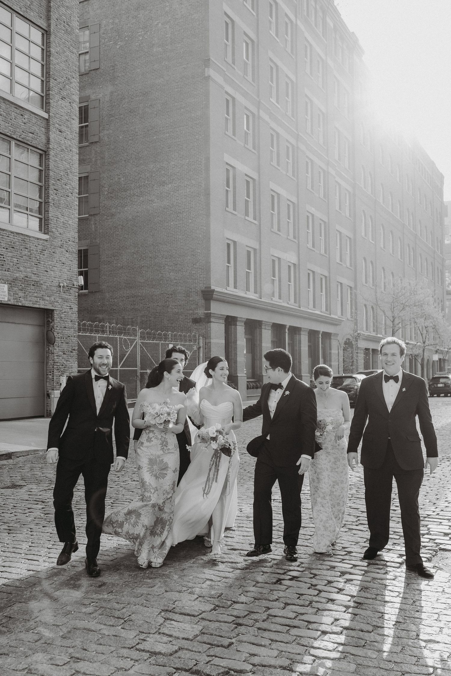 Black and white photo of wedding party walking on wet cobblestone street in NYC.