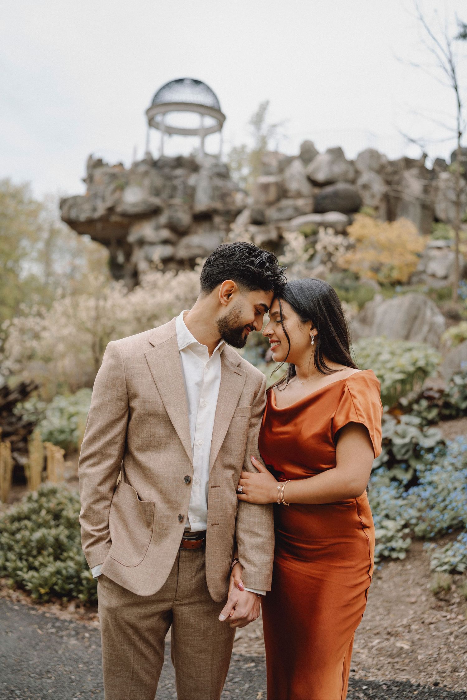 Couple touching foreheads tenderly with rocky landscape and glass dome gazebo structure in background.