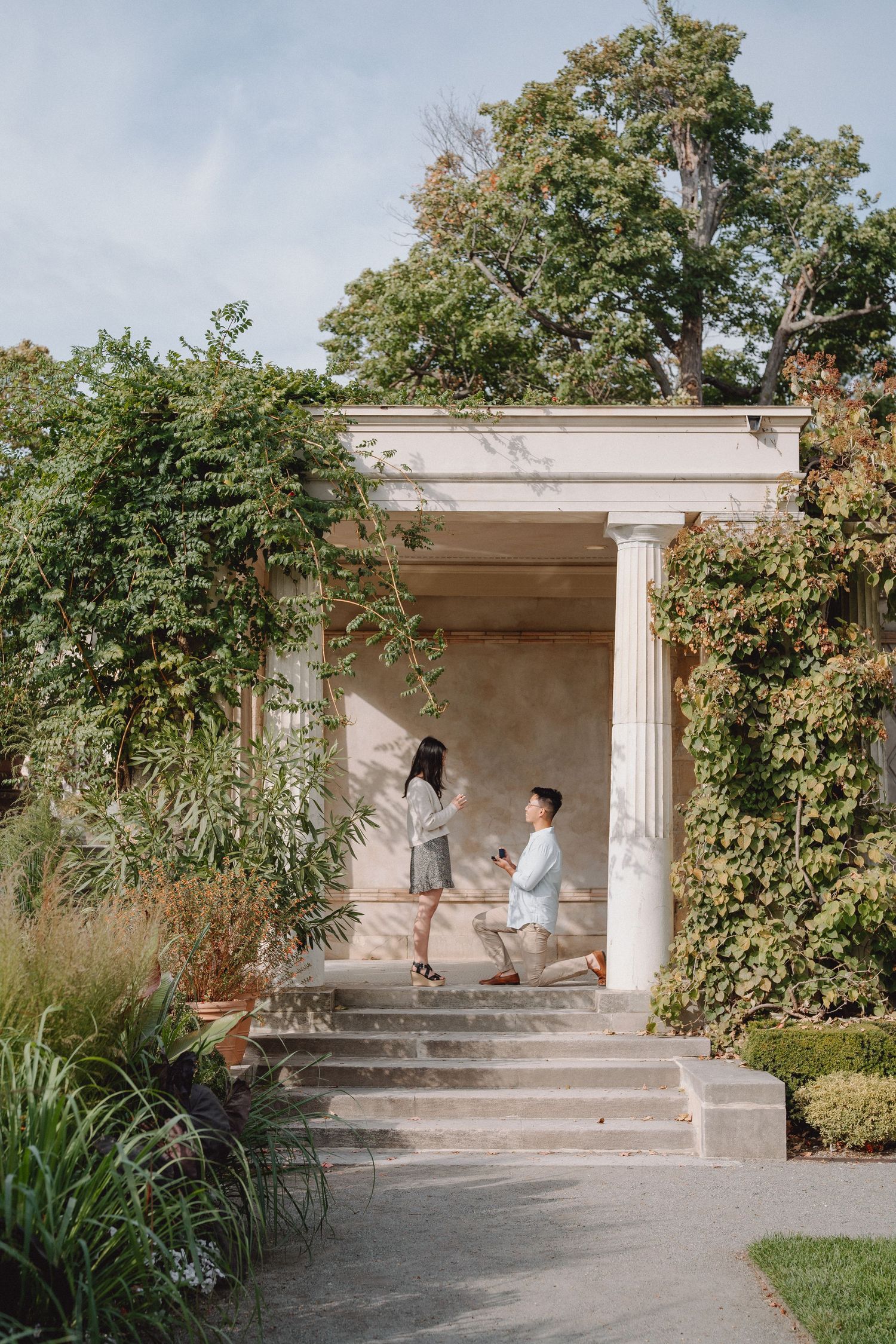 Outdoor marriage proposal on steps of classical pavilion surrounded by lush garden greenery.