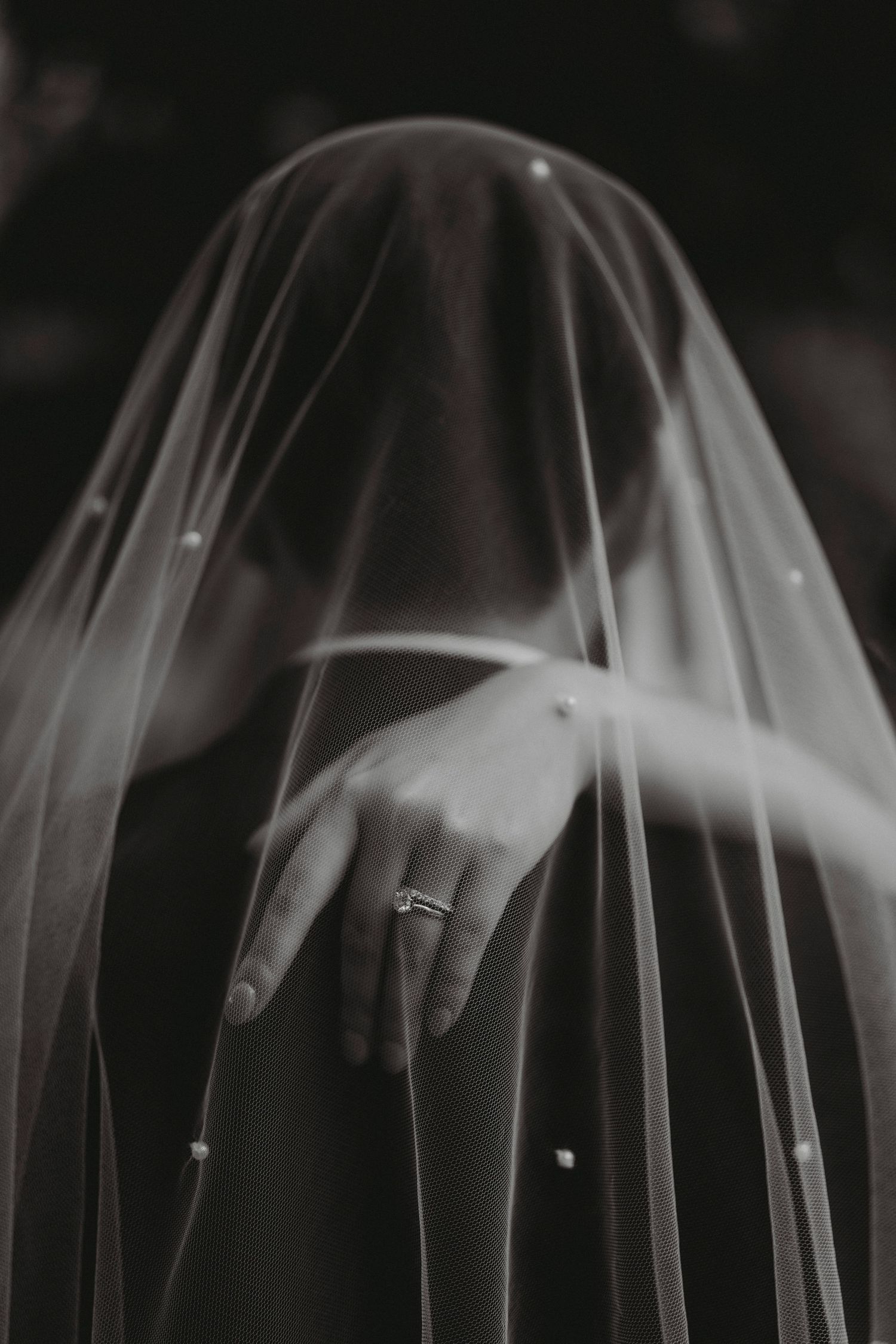 Close-up black and white shot of bride's hand with engagement ring beneath pearl-adorned veil.