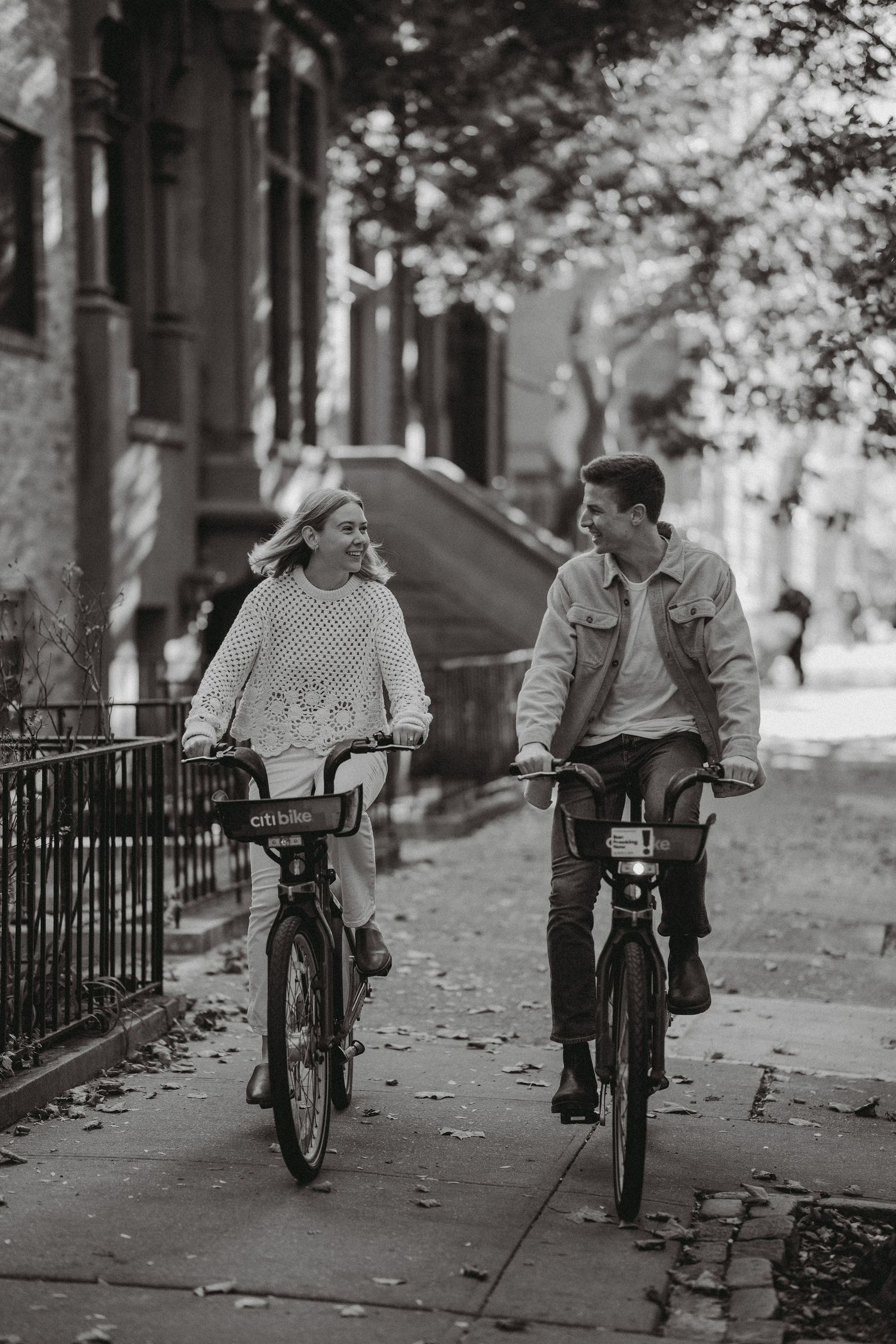 Black and white photo of a smiling couple riding Citi Bikes together on a tree-lined urban sidewalk.