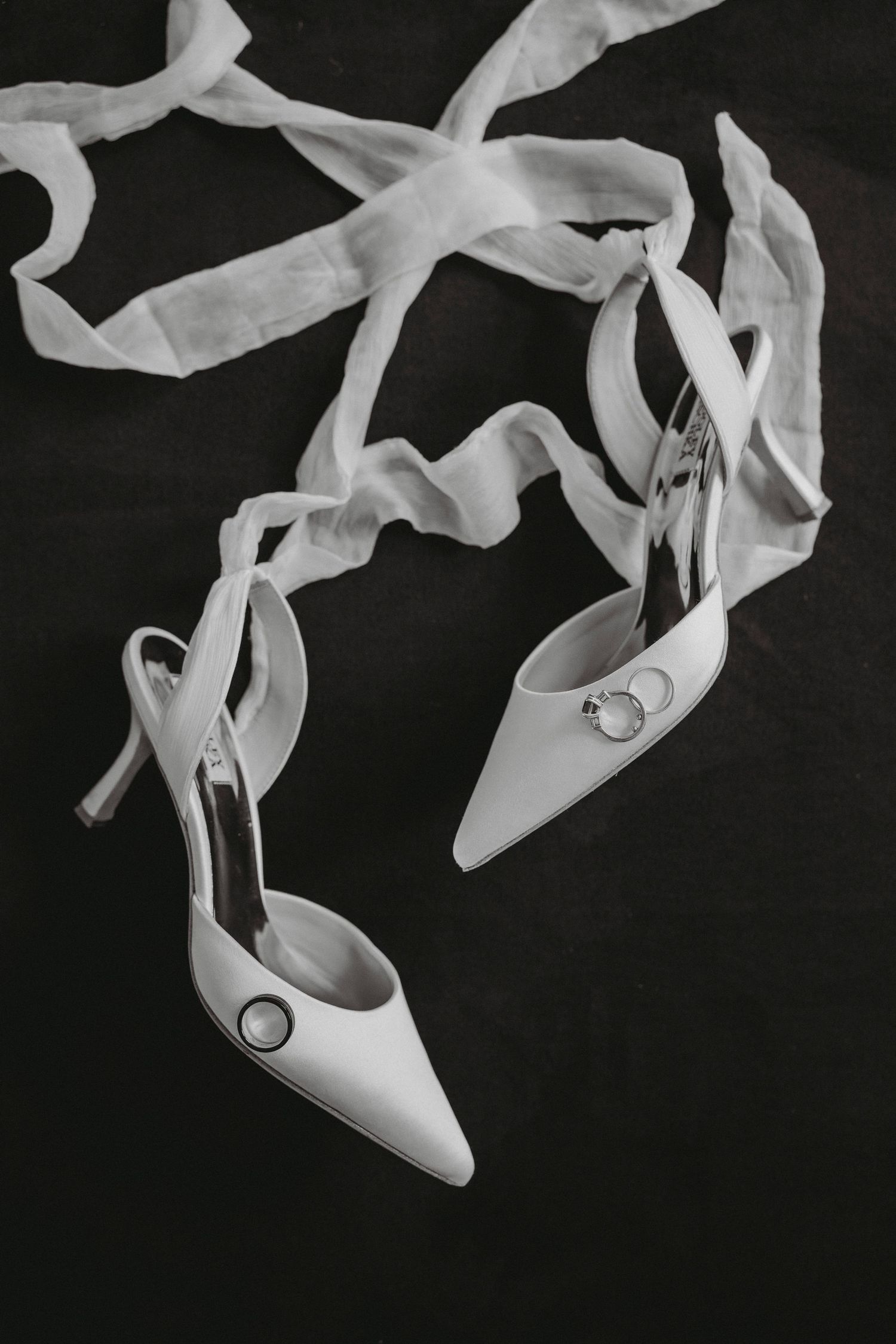 Black and white flat lay of elegant pointed-toe bridal heels with ribbon and diamond earrings.