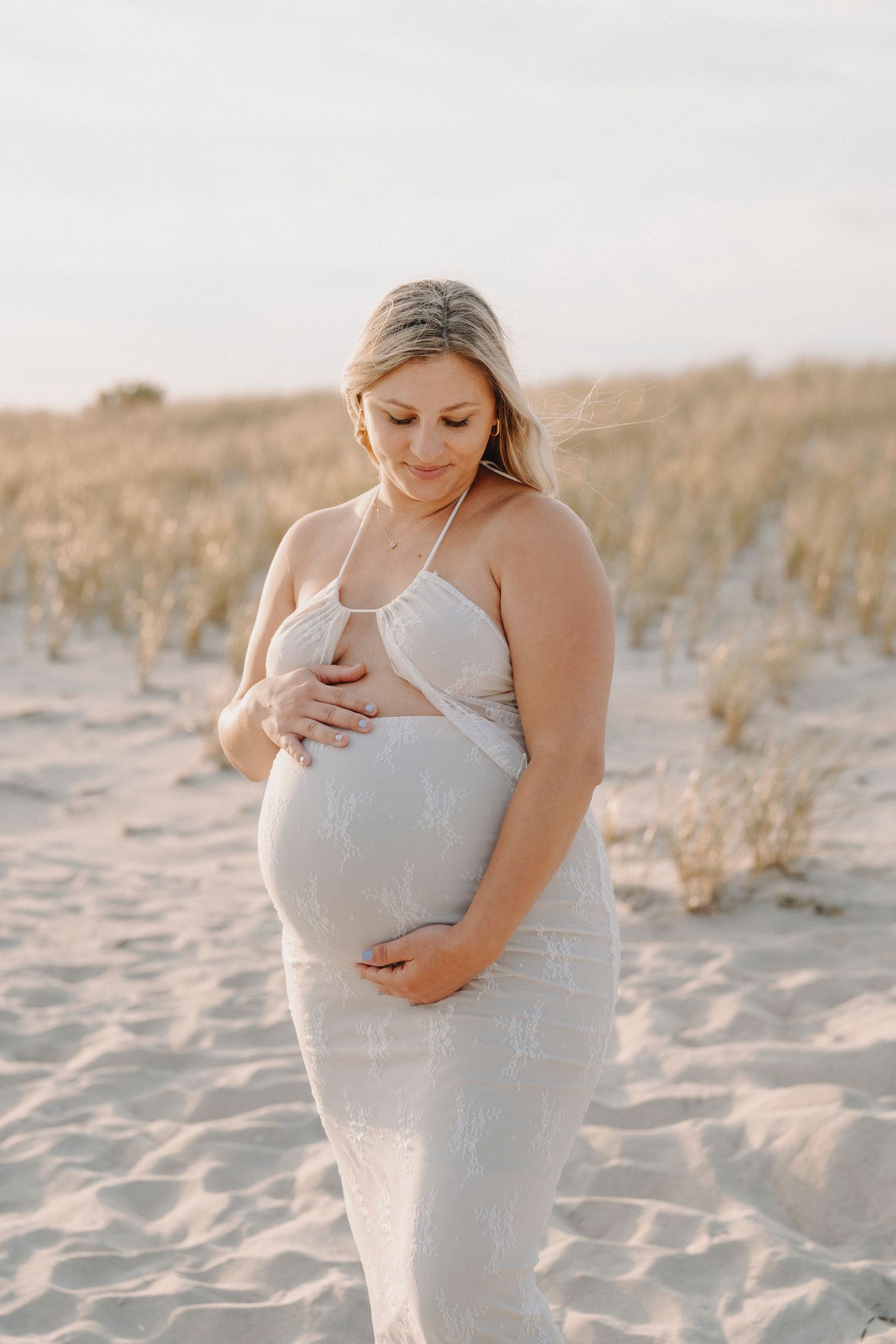 Pregnant blonde woman in white lace dress cradling belly on sandy beach at sunset.