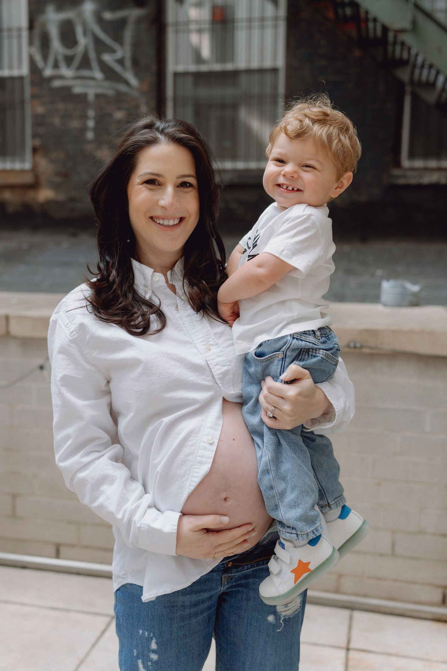 Smiling pregnant woman in white shirt holding toddler boy outdoors, showing baby bump.