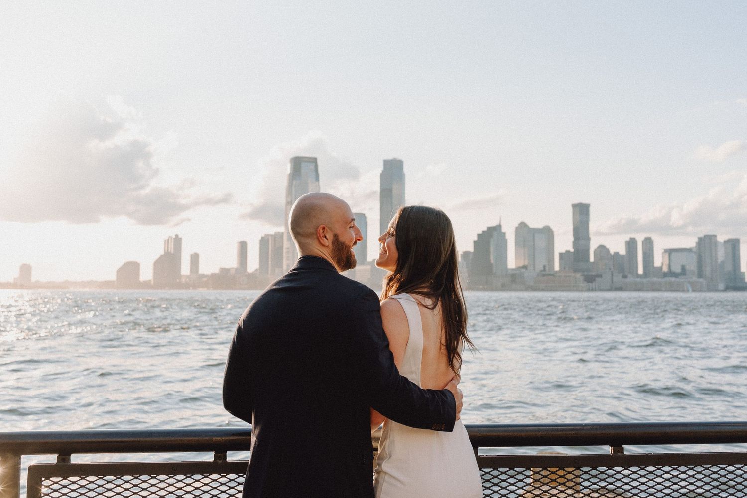 Couple embracing with the Jersey City skyline and Hudson River at sunset behind them.