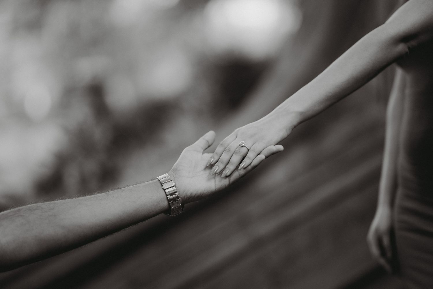 A close-up black and white photo of two hands gently touching, showcasing a delicate engagement ring.