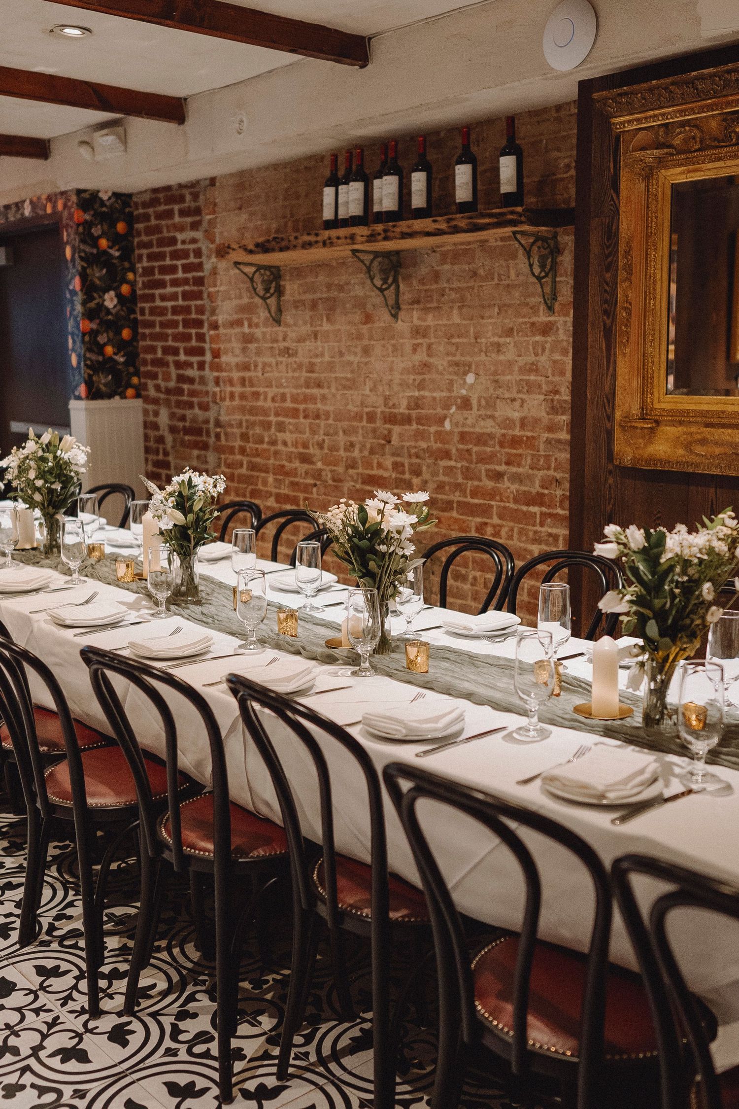 Long wedding reception table in rustic brick-walled venue with bentwood chairs and white floral arrangements.