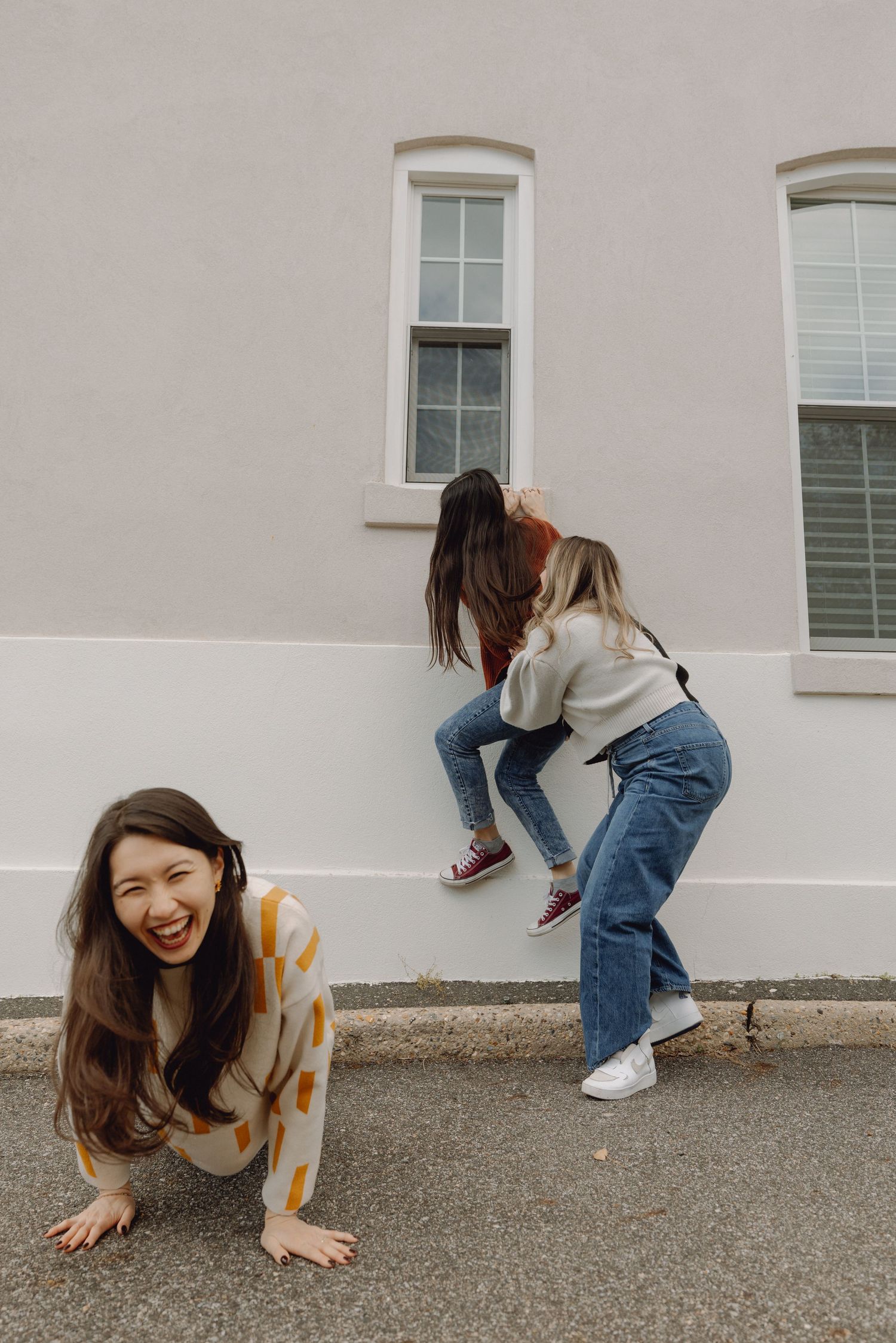 Three young women laughing and playing outside a building, one crawling on ground while two climb a wall.