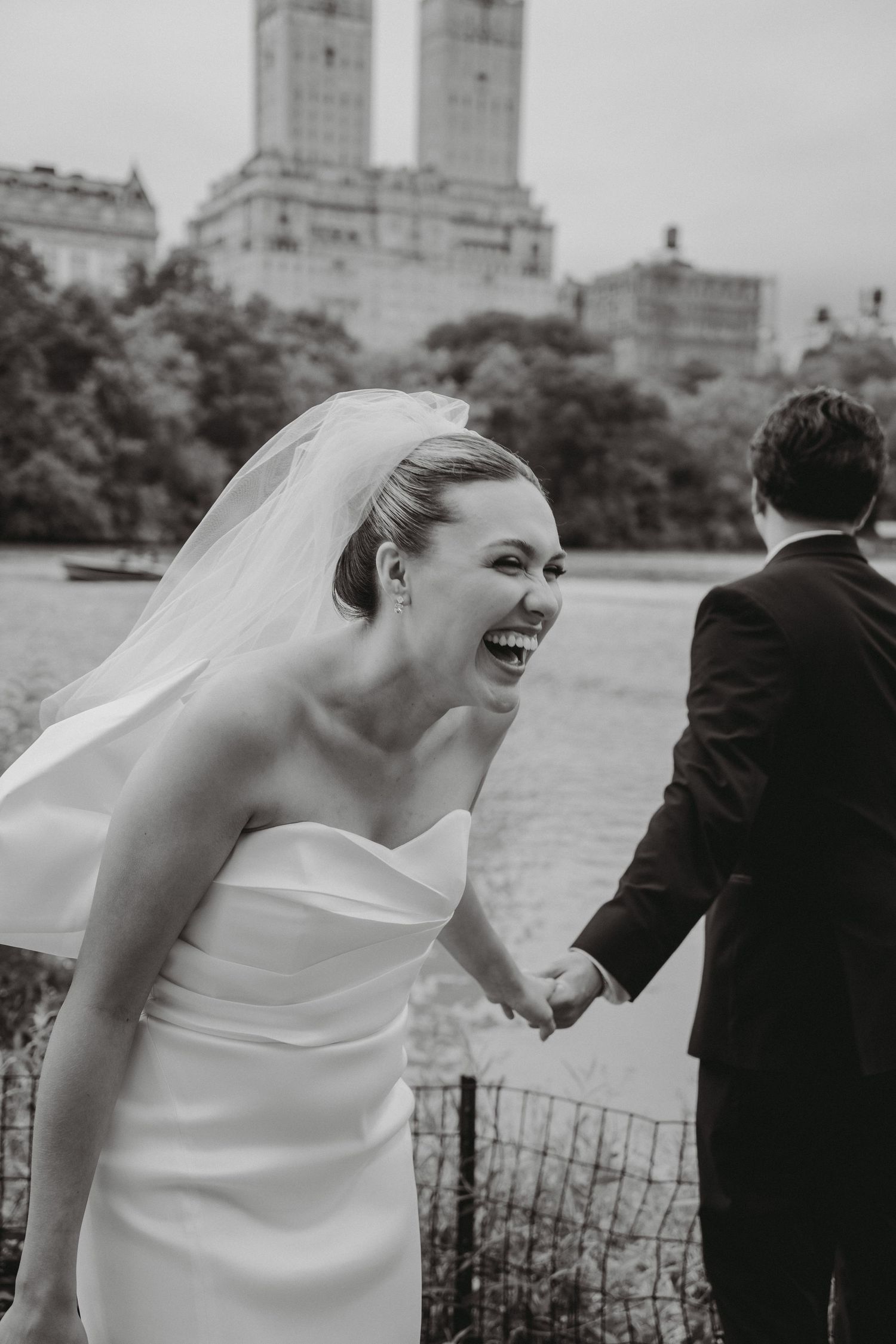 Black and white photo of joyful bride laughing while holding grooms hand near Central Park waterfront.