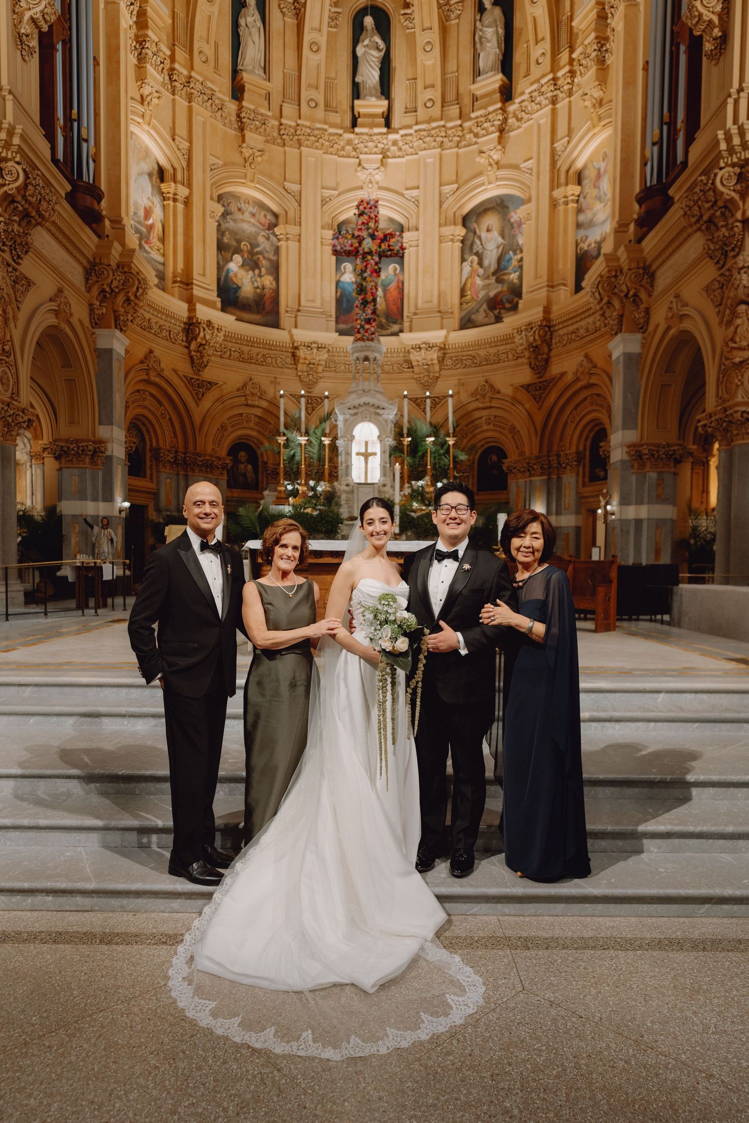 Bride and groom pose with four family members before elaborate gilded church altar.