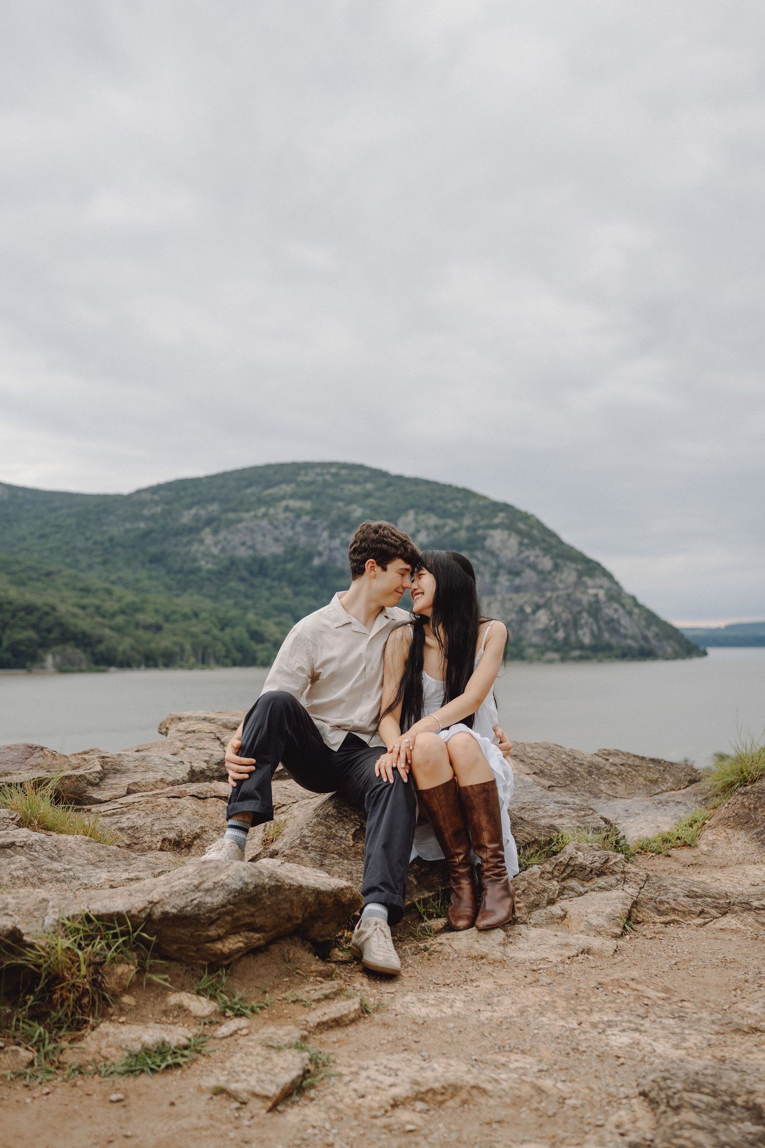 Couple sitting close on a rocky overlook, foreheads touching, with a calm river and forested mountain behind them under a cloudy sky.