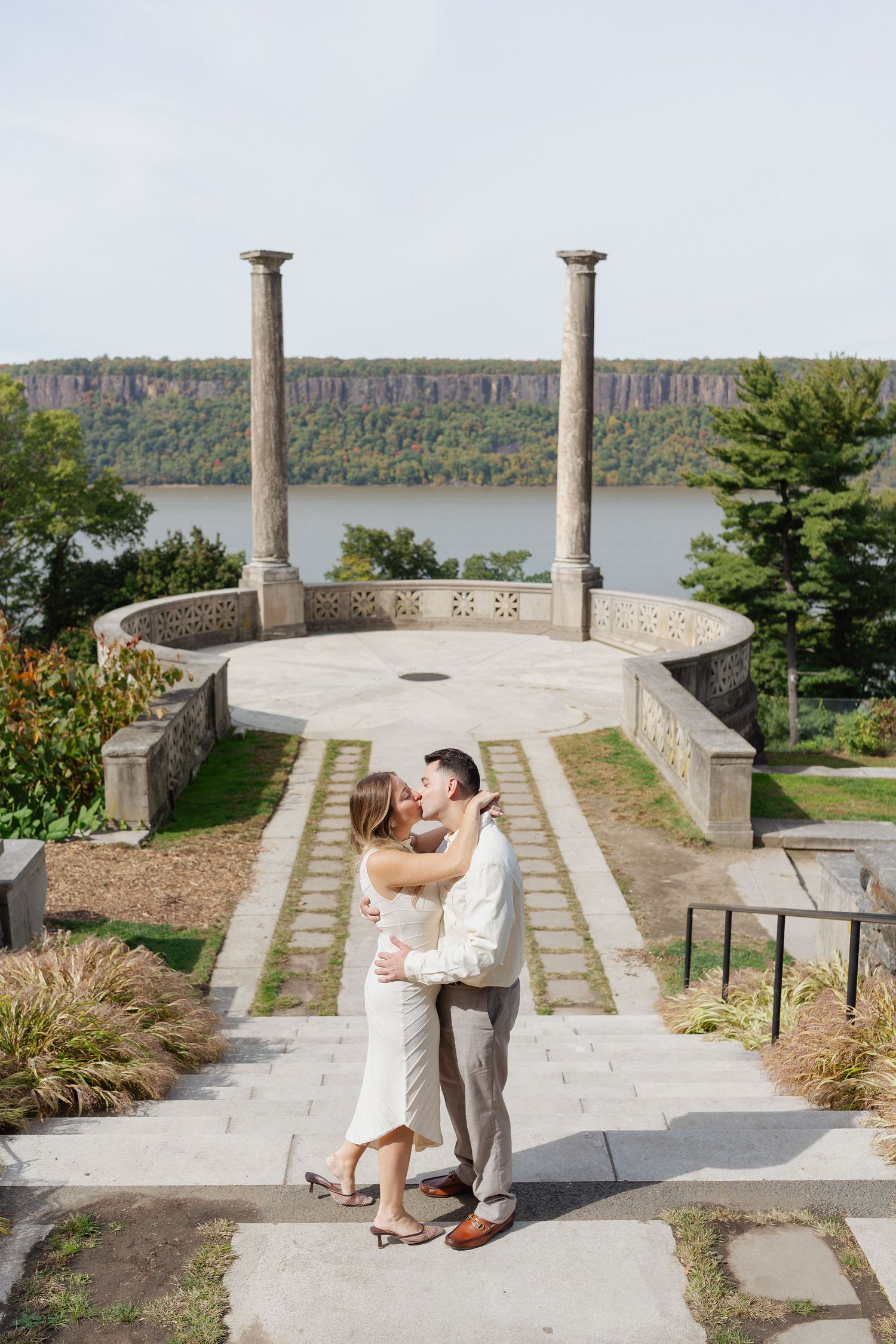 Couple embracing and kissing on stone steps in front of a circular terrace with two columns overlooking a river and cliffs.