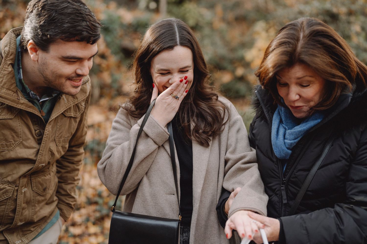 Smiling woman covers her mouth with her hand, moved to tears, while a man and another woman look on in an autumn park.