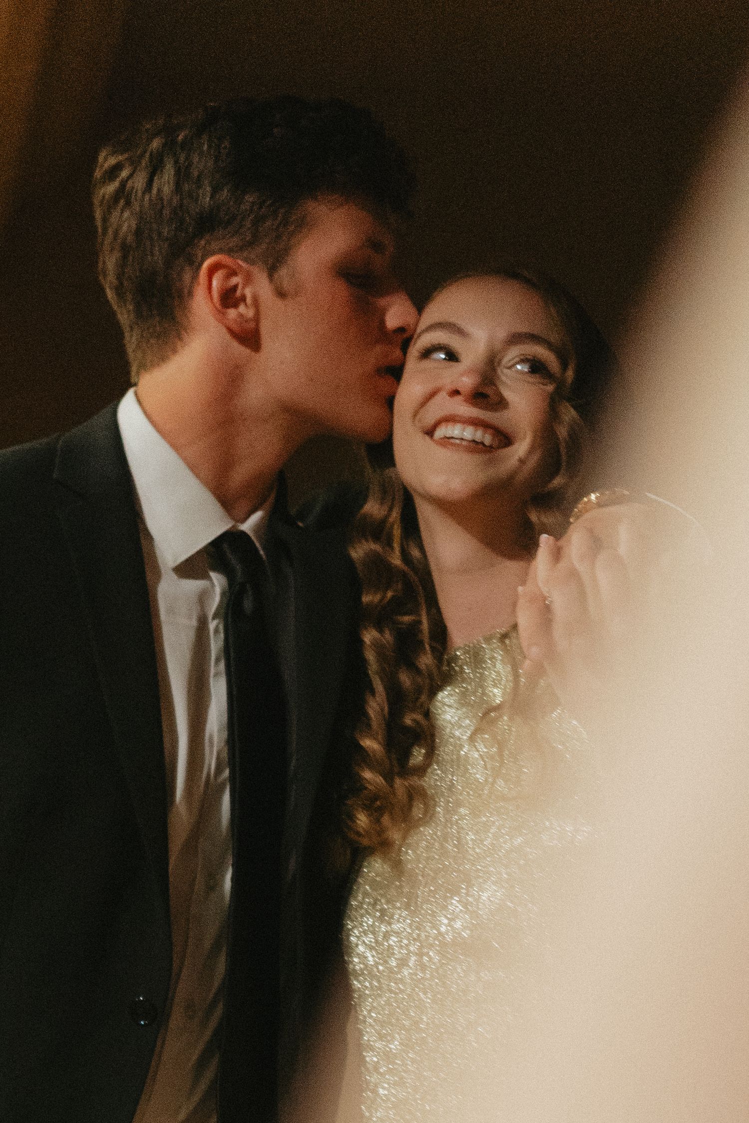 A young couple in formal attire shares a tender moment, with the man kissing the smiling woman on the cheek.