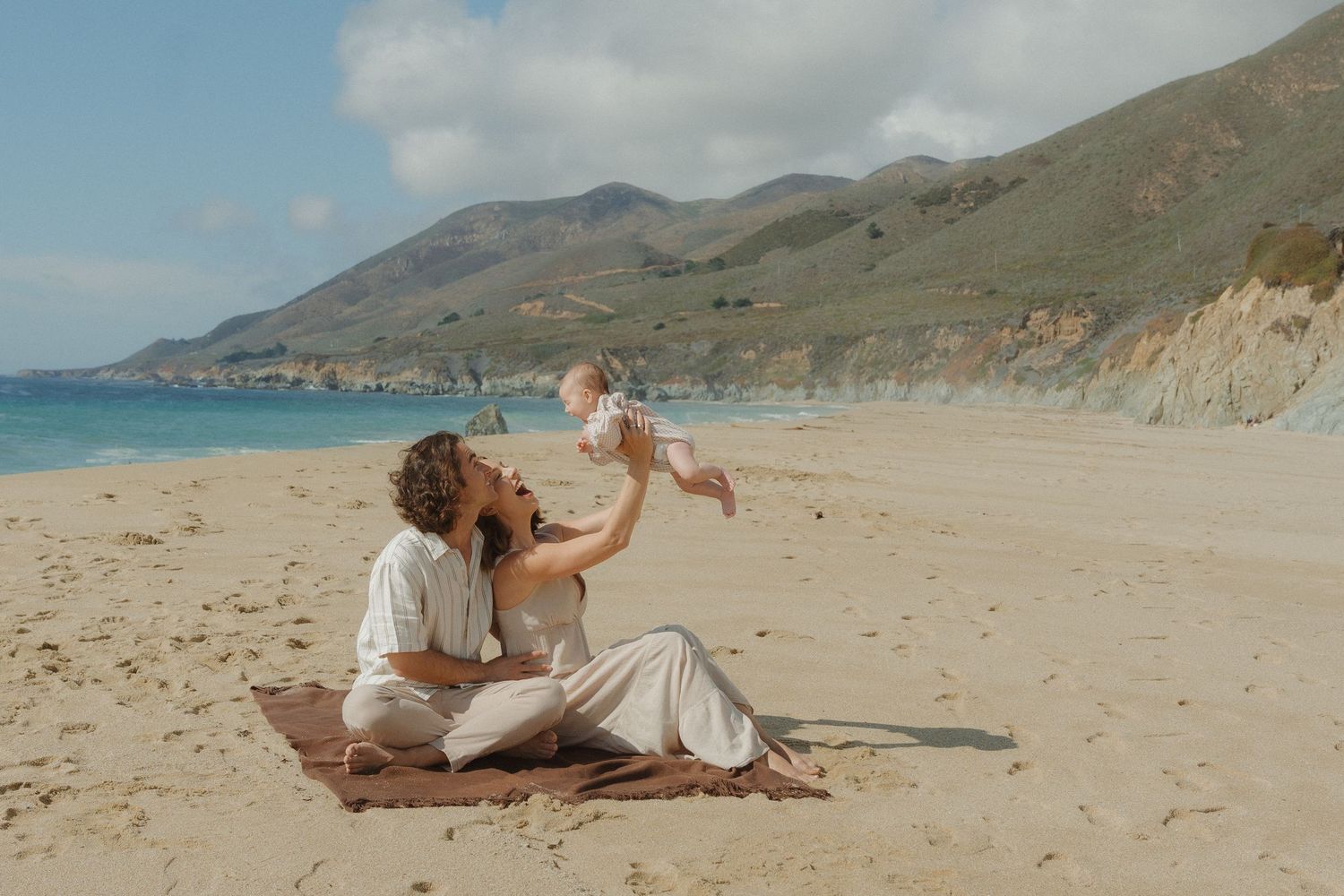Mother lifting her baby joyfully while sitting on a beach blanket with scenic coastal mountains at Carmel, CA.