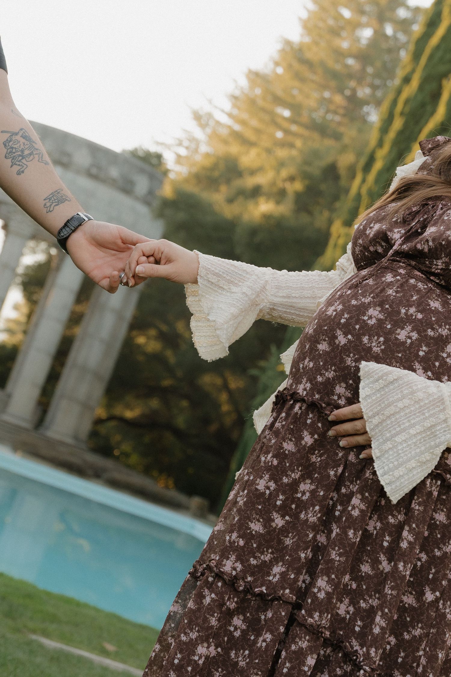 Pregnant woman in floral brown dress holding hands with tattooed partner near a garden pool at Redwood City.