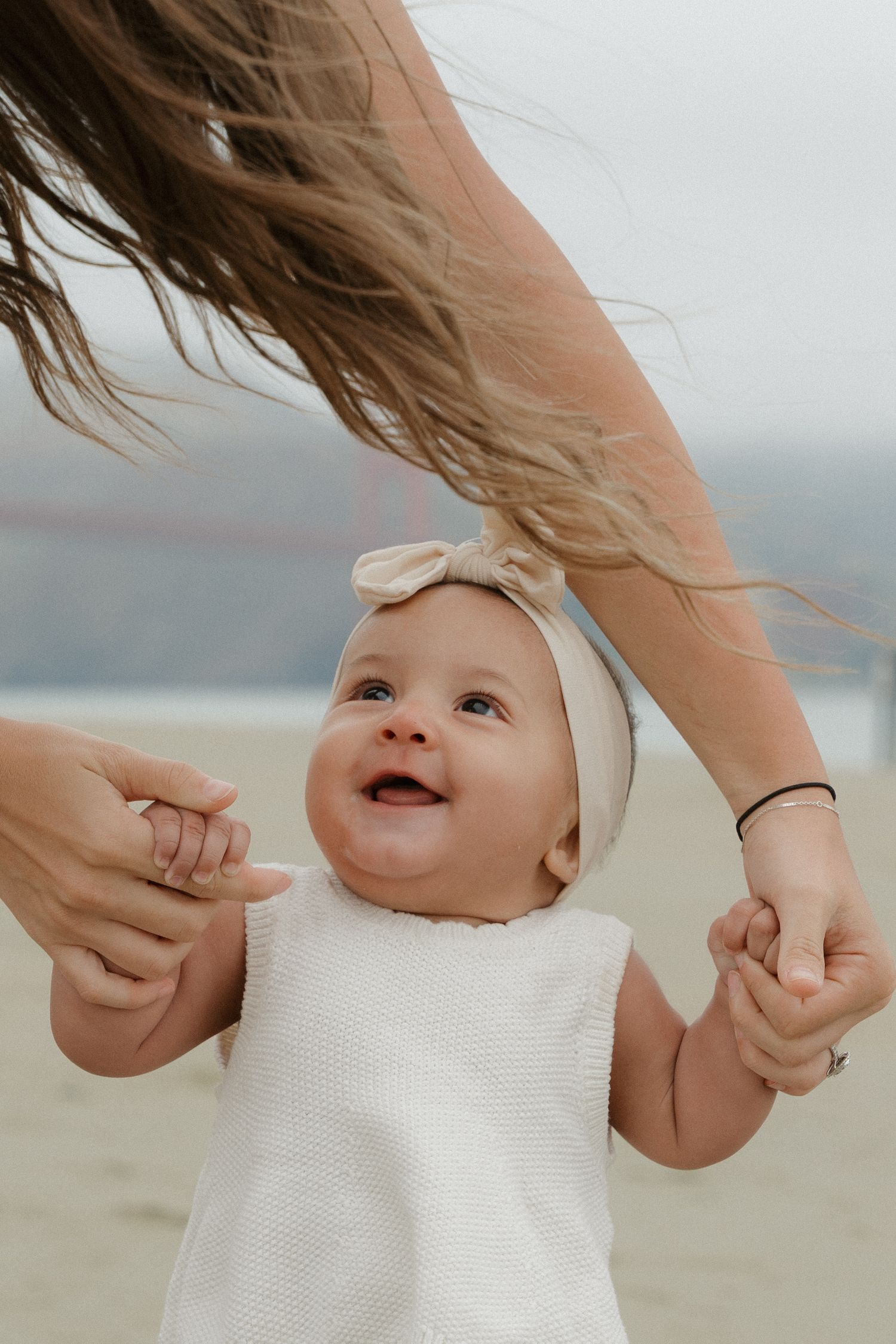 A joyful baby in a white outfit and bow headband holds a mother's hands on a beach near the Golden Gate Bridge.