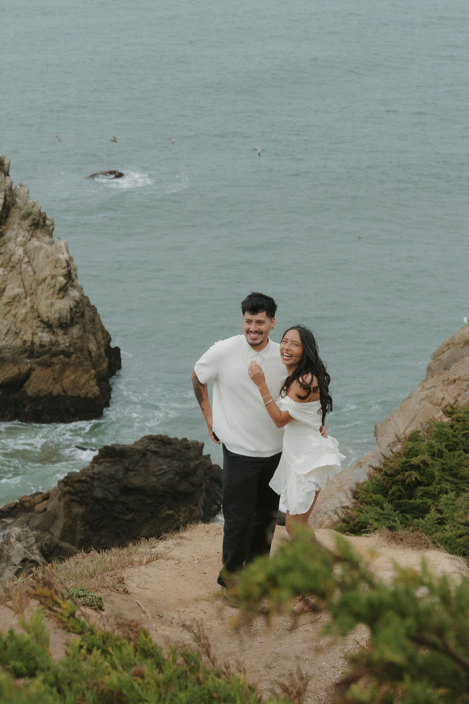 Happy engaged couple hugging and smiling on rocky coastal cliffs above turquoise ocean waters at Lands End, CA.