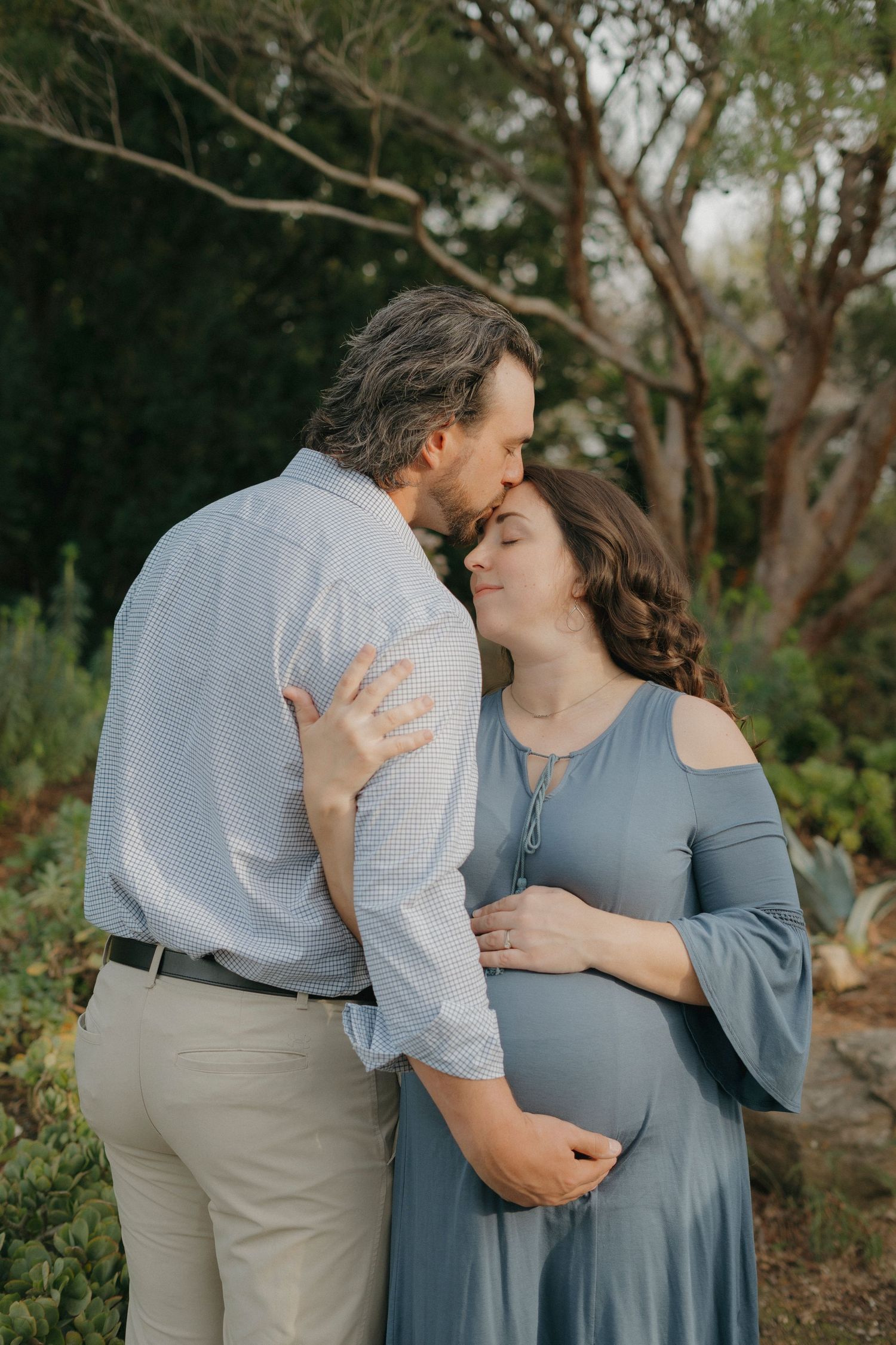 Expectant couple sharing a tender kiss outdoors surrounded by lush garden greenery in Walnut Creek, CA.