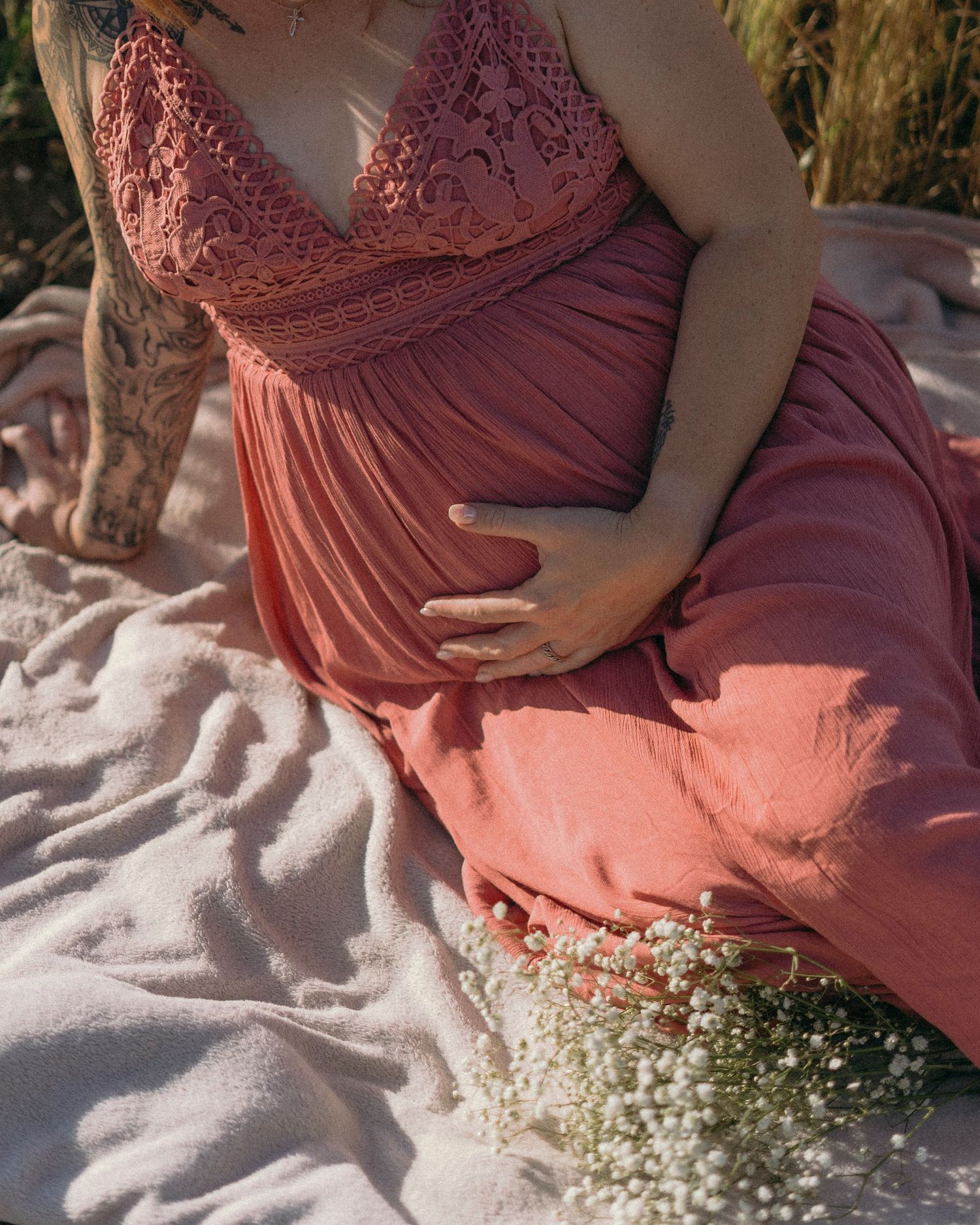 Pregnant woman in a rose pink lace materniy dress sitting outdoors holding her baby bump with baby's breath flowers in Woodland, CA.