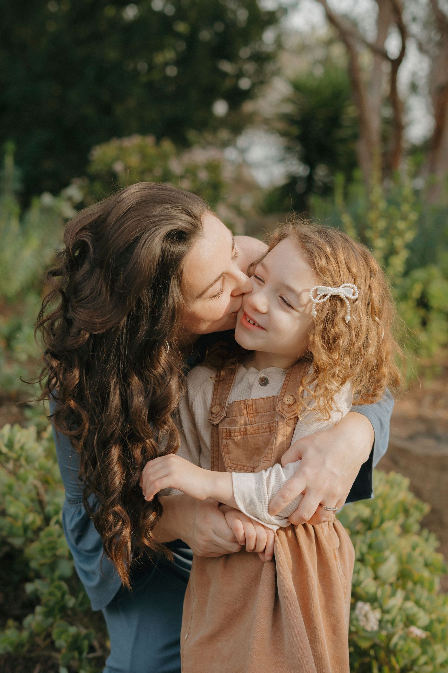 Mother with long curly hair kissing her smiling red-haired daughter in a sunlit garden at Walnut Creek, CA.