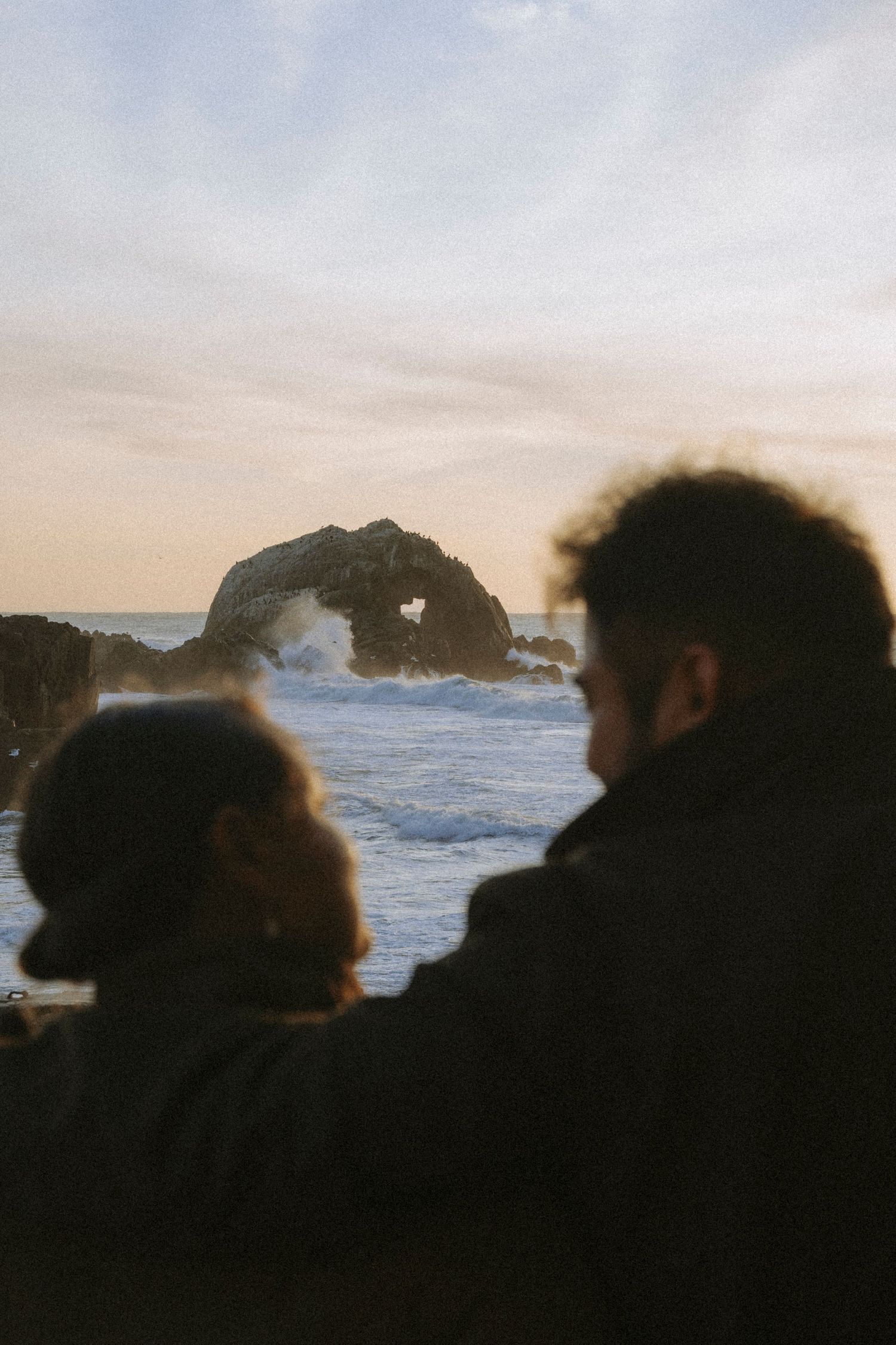Couple silhouetted against a dramatic coastal sunset with rocky sea arch and crashing waves in the background at Lands End.