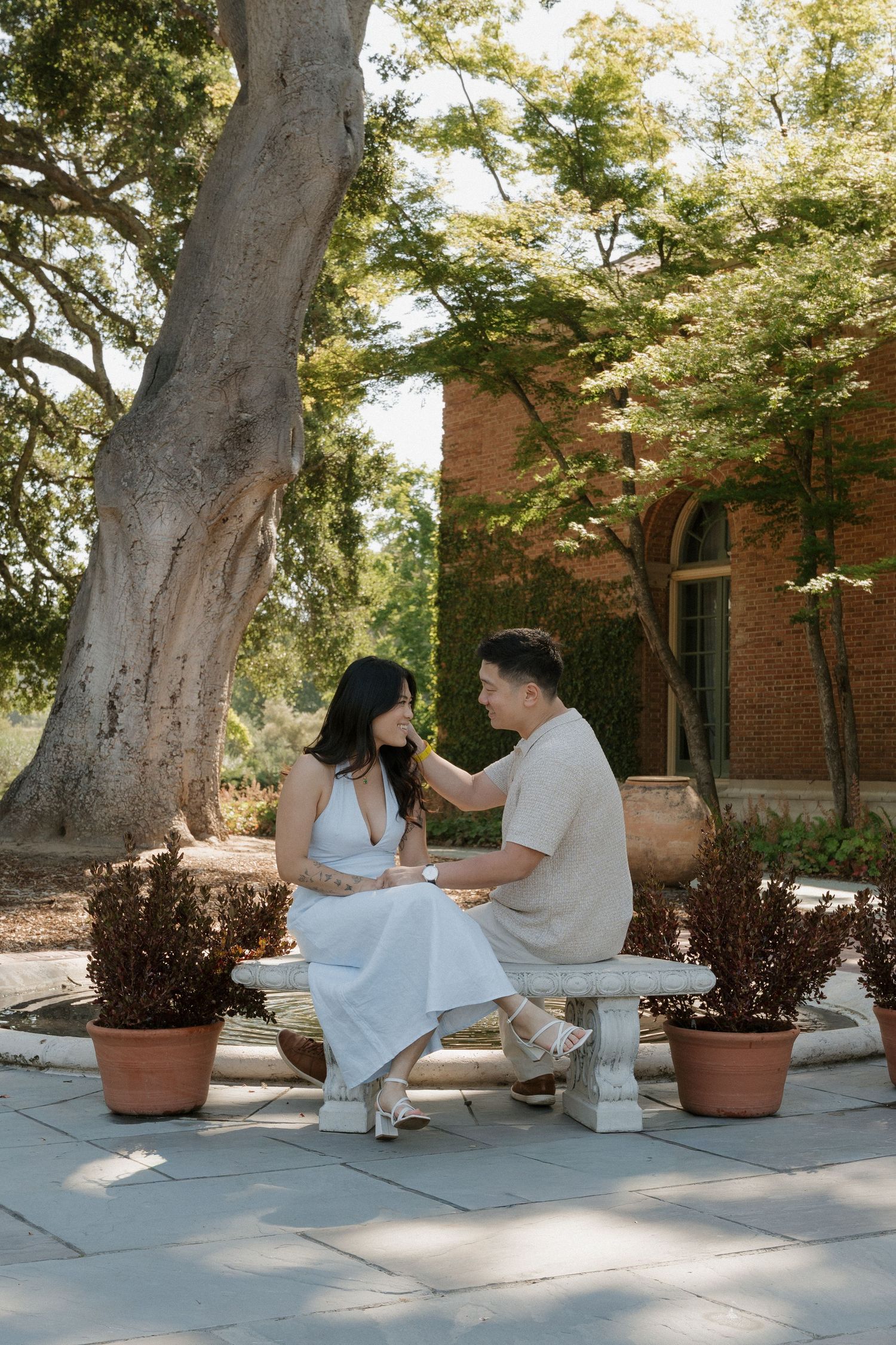 Couple sitting on a stone bench in a sunlit garden, surrounded by potted plants and lush trees near a brick building.
