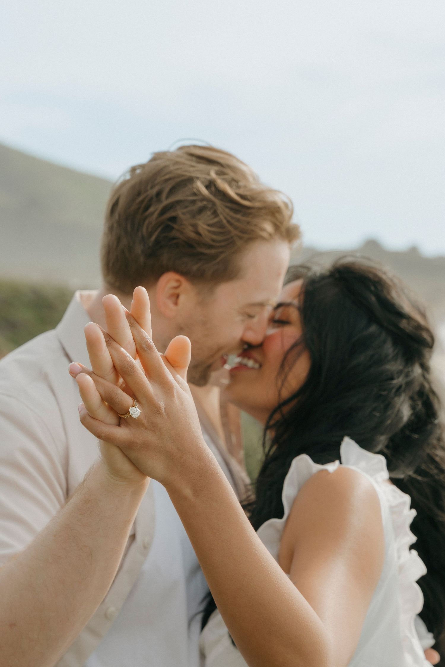 Newly engaged couple kissing while showing off diamond engagement ring with mountains behind in Carmel, CA.