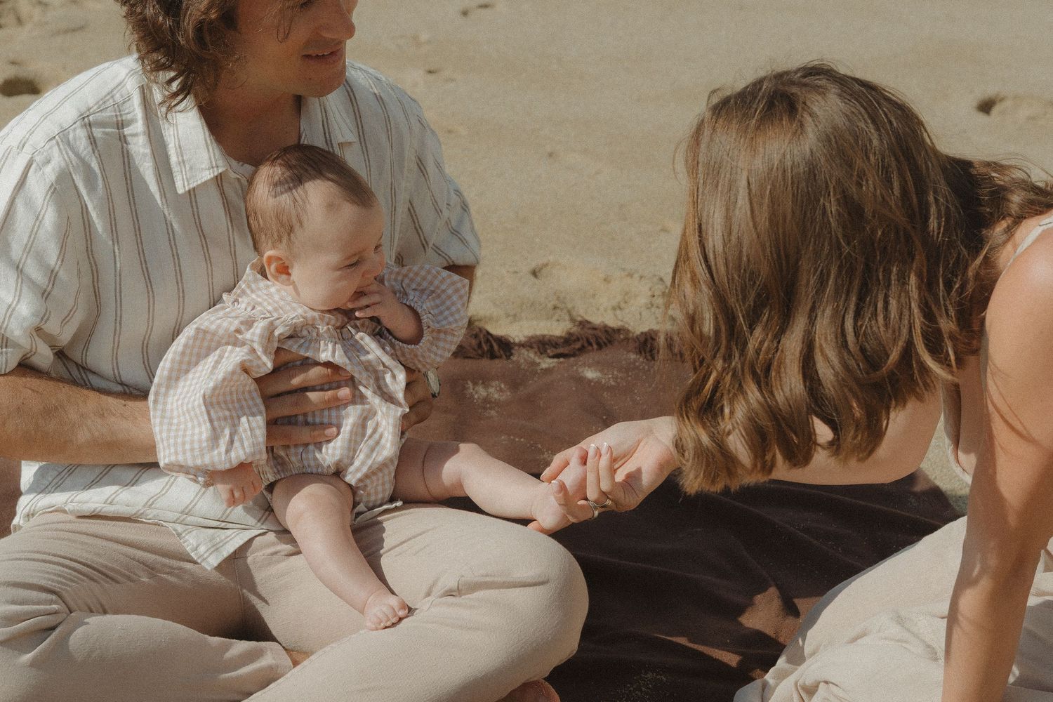 Young parents sitting on a sandy beach with their smiling baby between them.