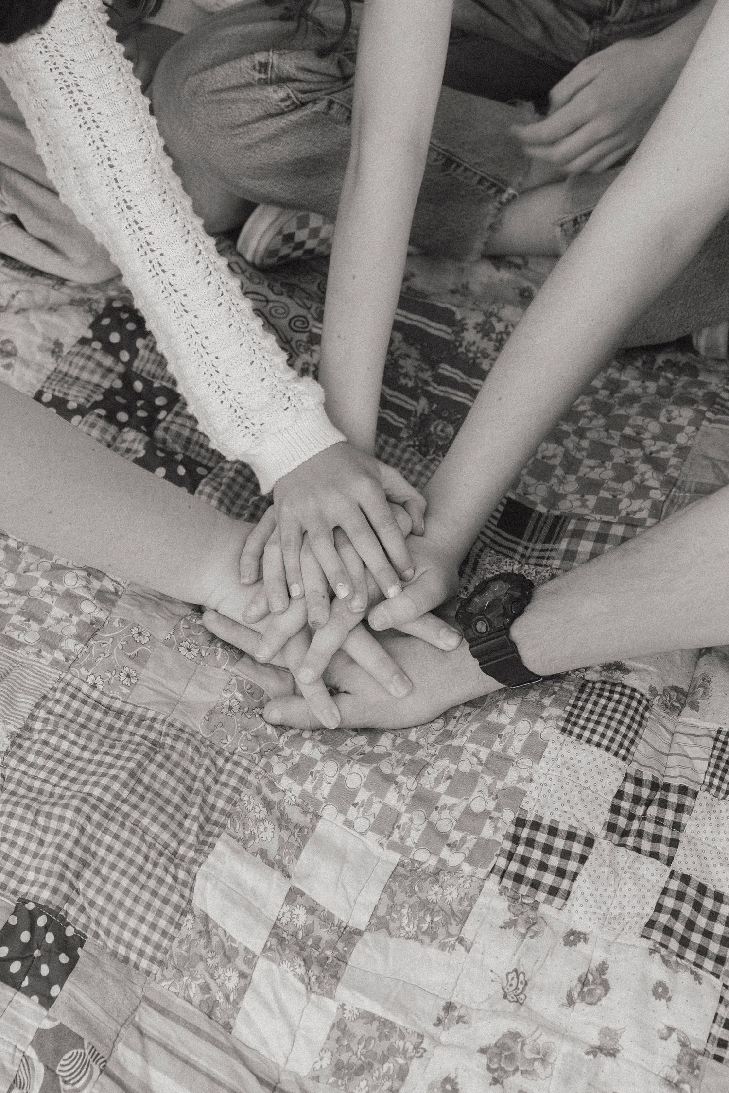 Black and white photo of multiple hands stacked together on a patchwork quilt in unity.