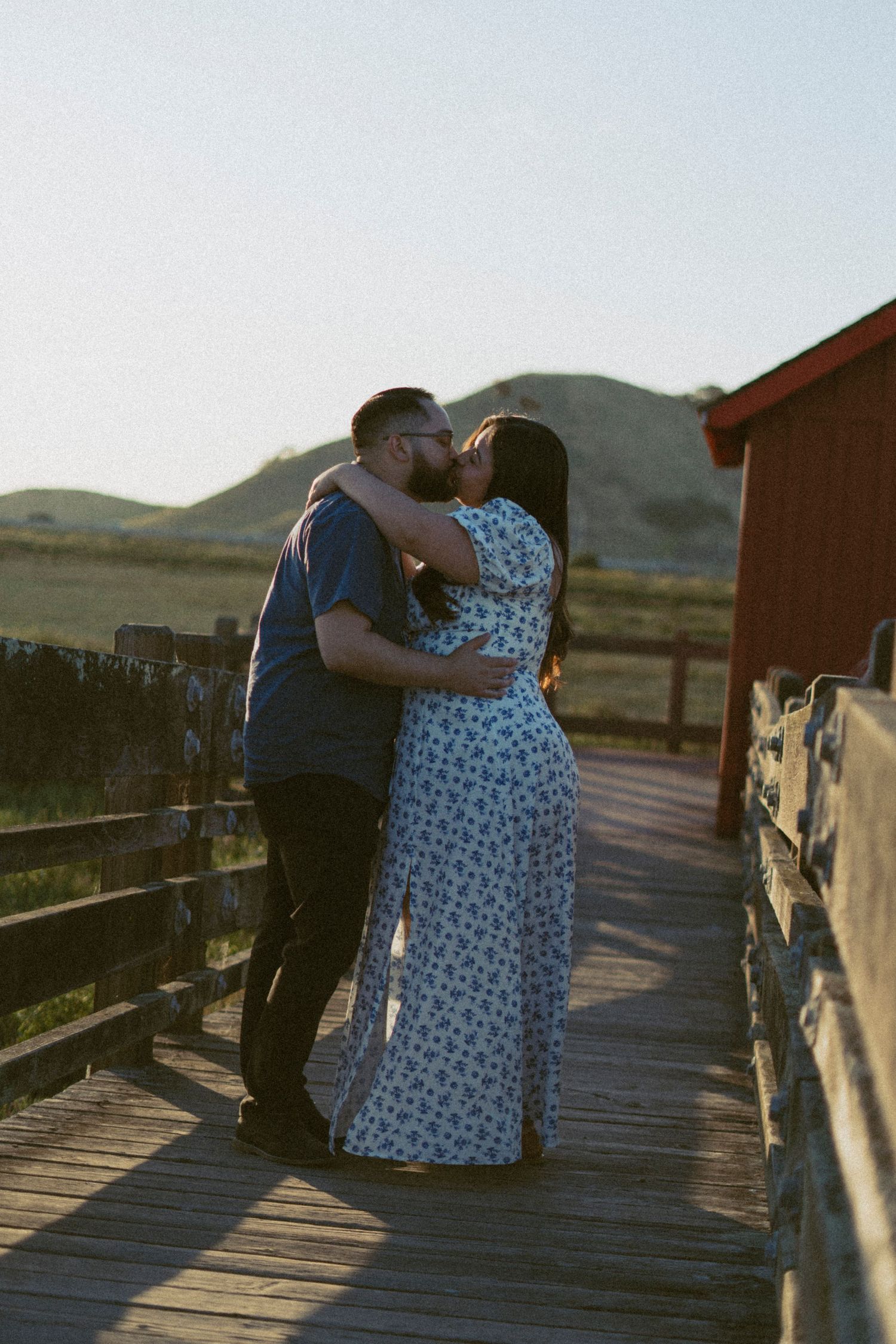 Couple sharing a romantic kiss on a wooden boardwalk at golden hour with rolling hills behind in Fremont, CA.
