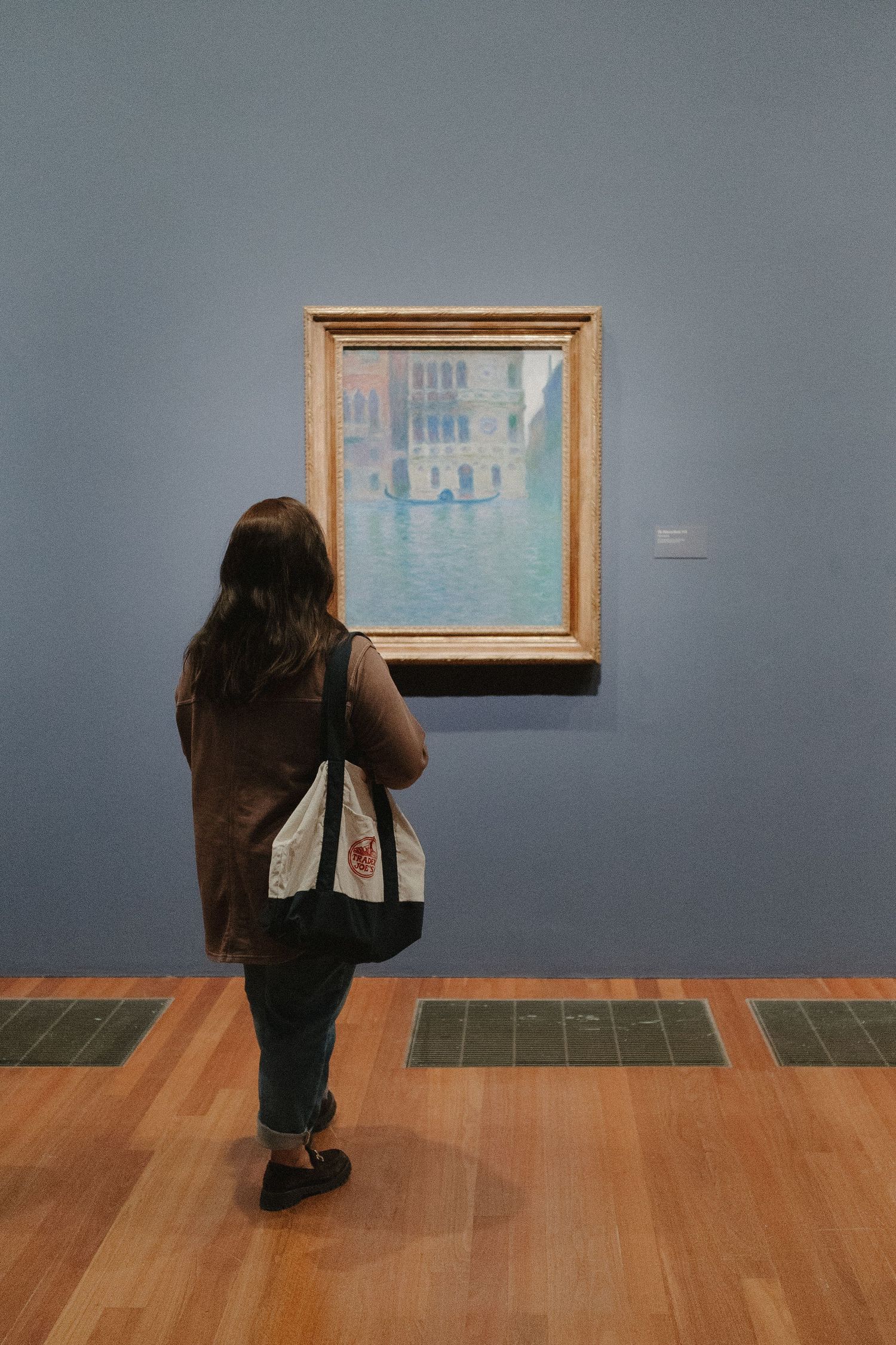 Woman with tote bag viewing a framed Venetian canal painting in a blue-walled art museum gallery.