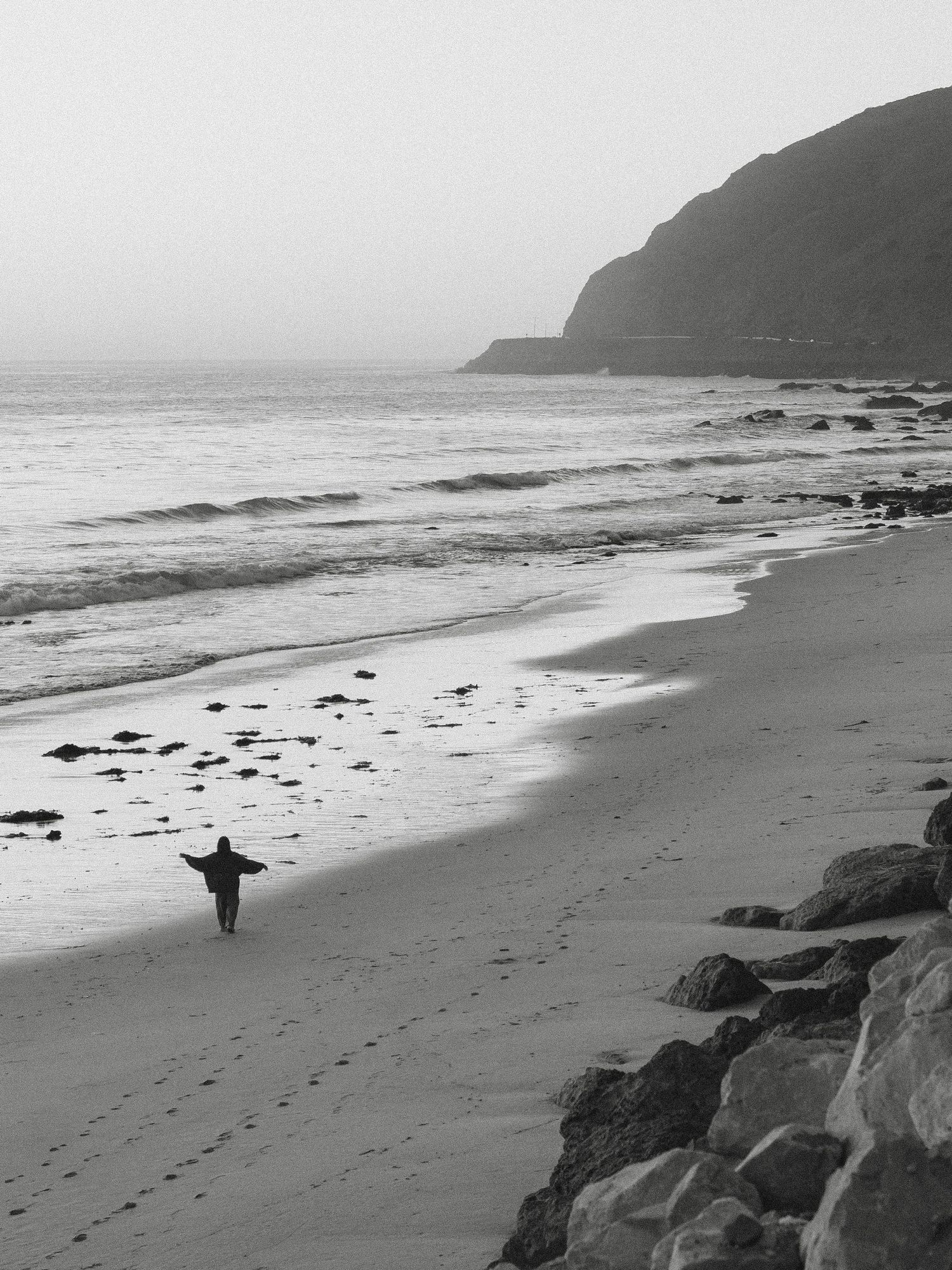 Black and white coastal landscape with lone figure standing on a misty beach near rocky cliffs.