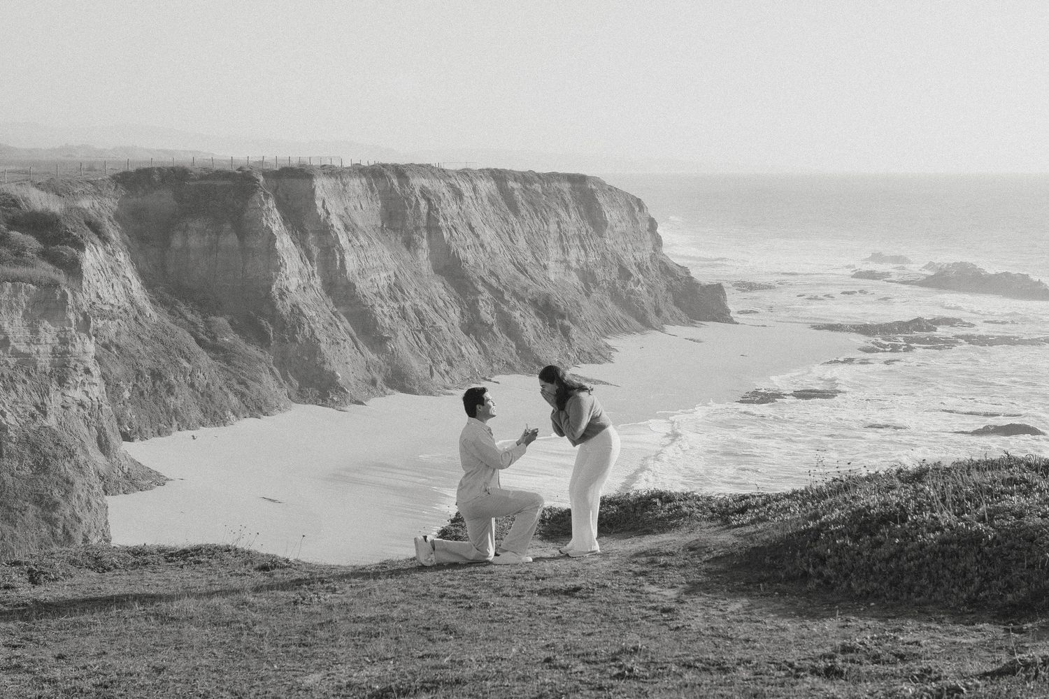 Black and white photo of a romantic marriage proposal on a coastal cliff overlooking the ocean and sandy beach.