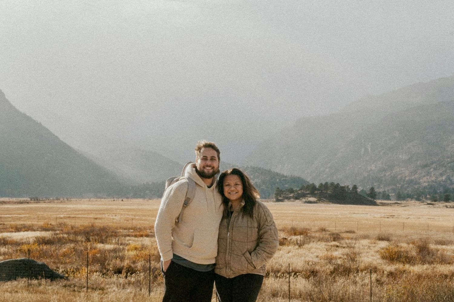 Smiling couple embracing in golden meadow with misty mountain range backdrop in autumn landscape.