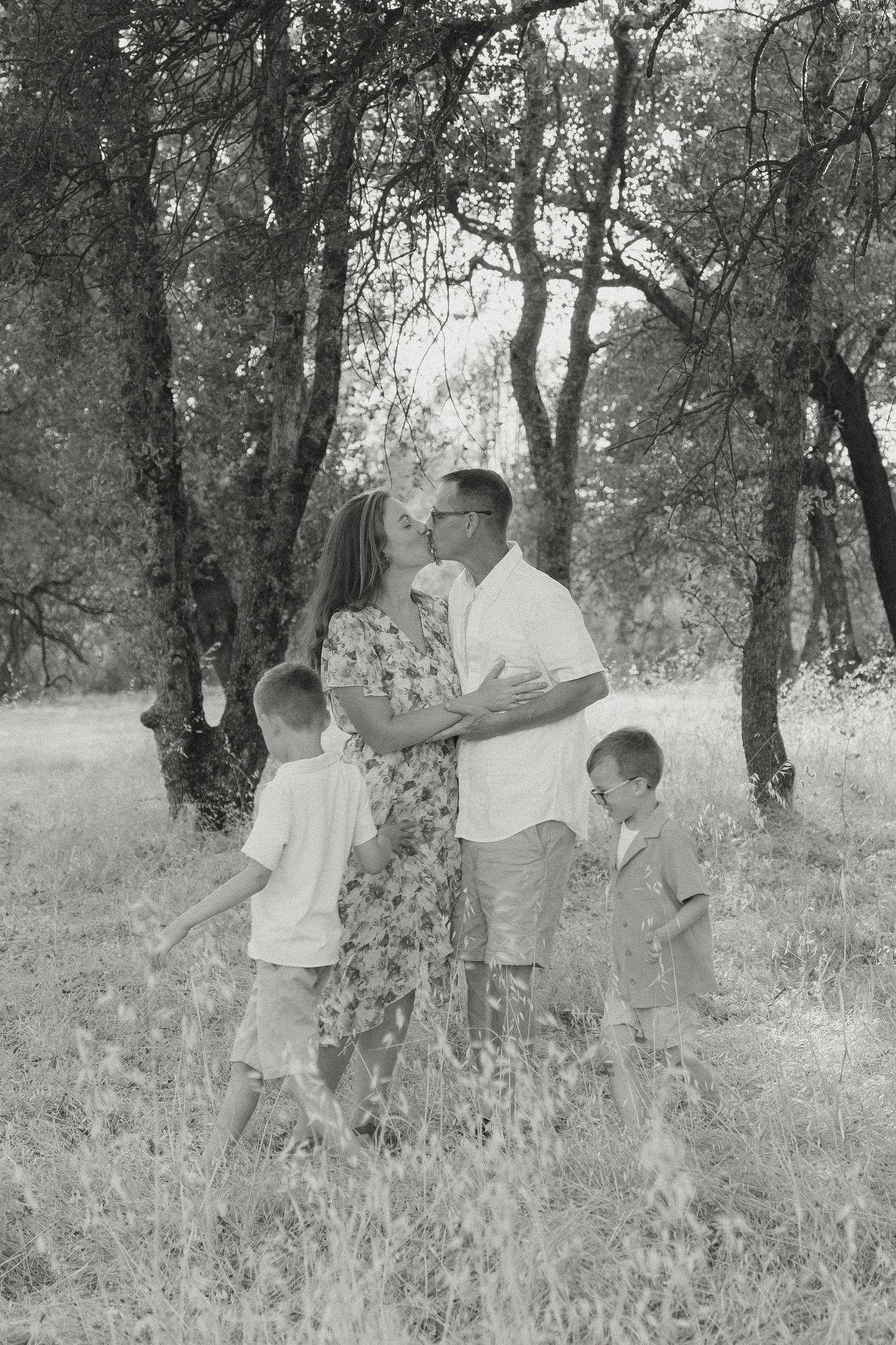 Black and white family portrait of parents kissing with two young sons in a wooded field in Marysville, CA.
