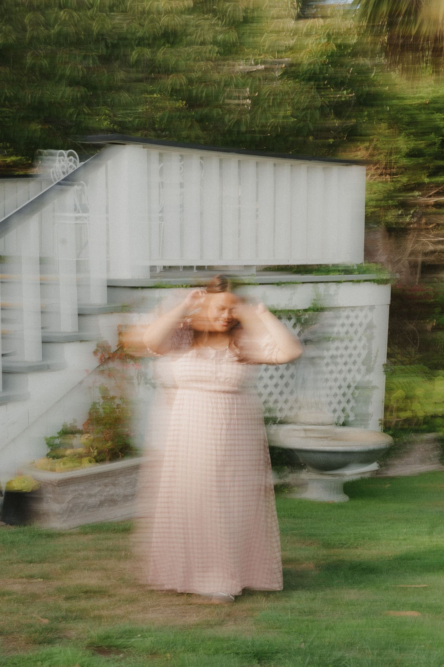 A blurred woman in a pink dress poses outdoors near white stairs and garden in a dreamy motion-effect photo.