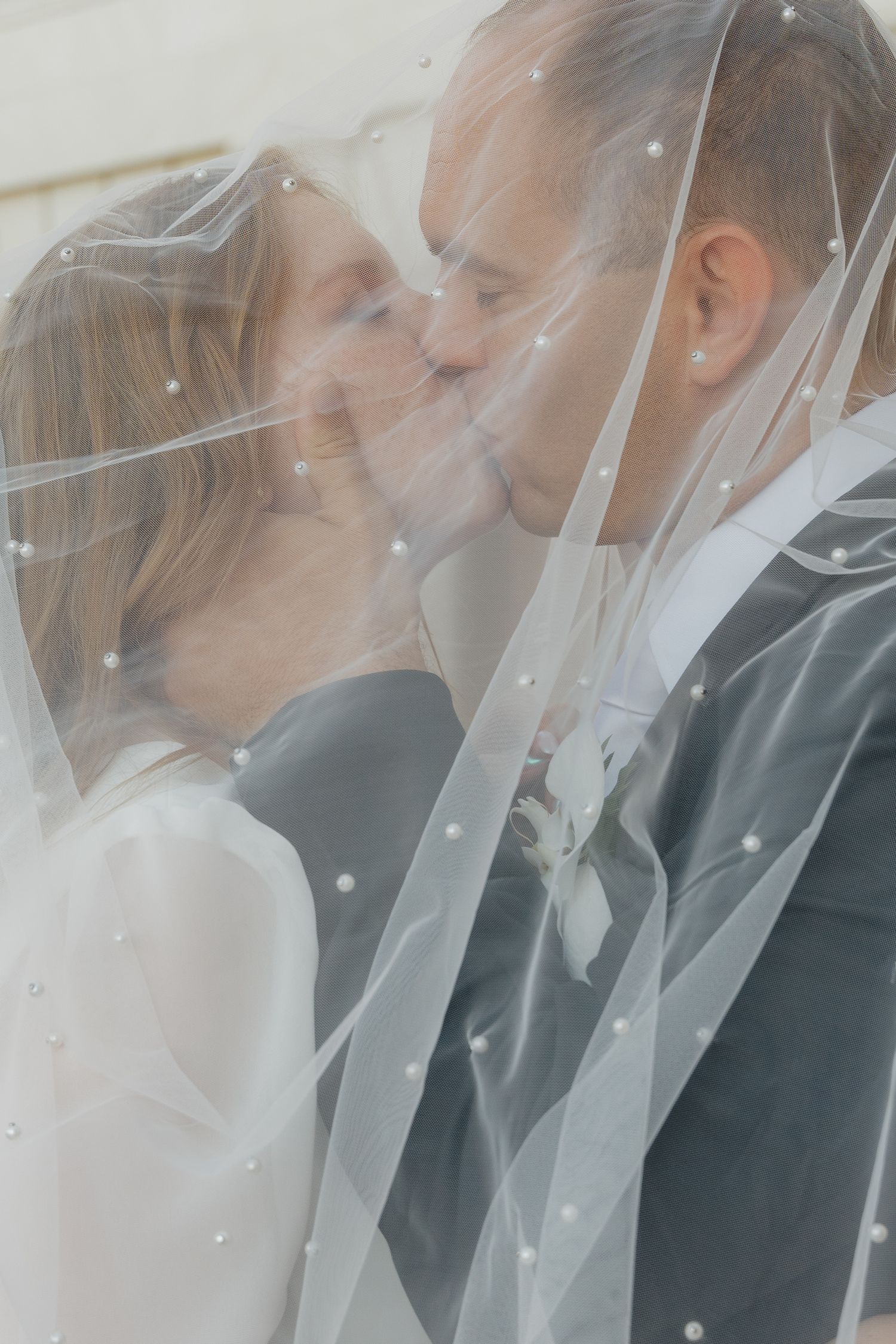 Bride and groom sharing a romantic kiss beneath a pearl-adorned wedding veil.