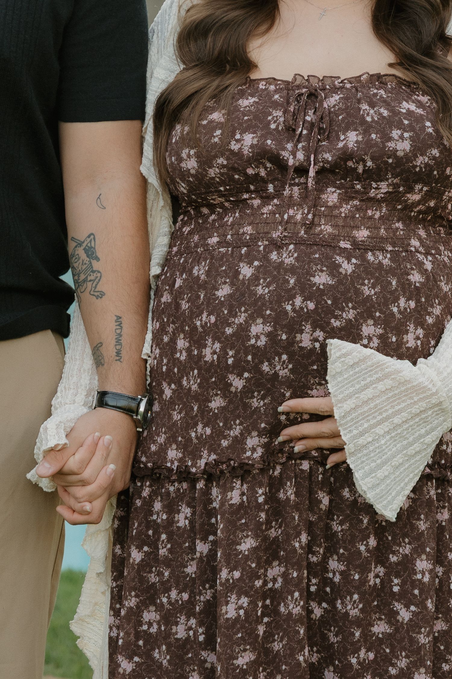 Pregnant woman in brown floral dress holds hands with tattooed partner during maternity photo session outdoors in Redwood City, CA