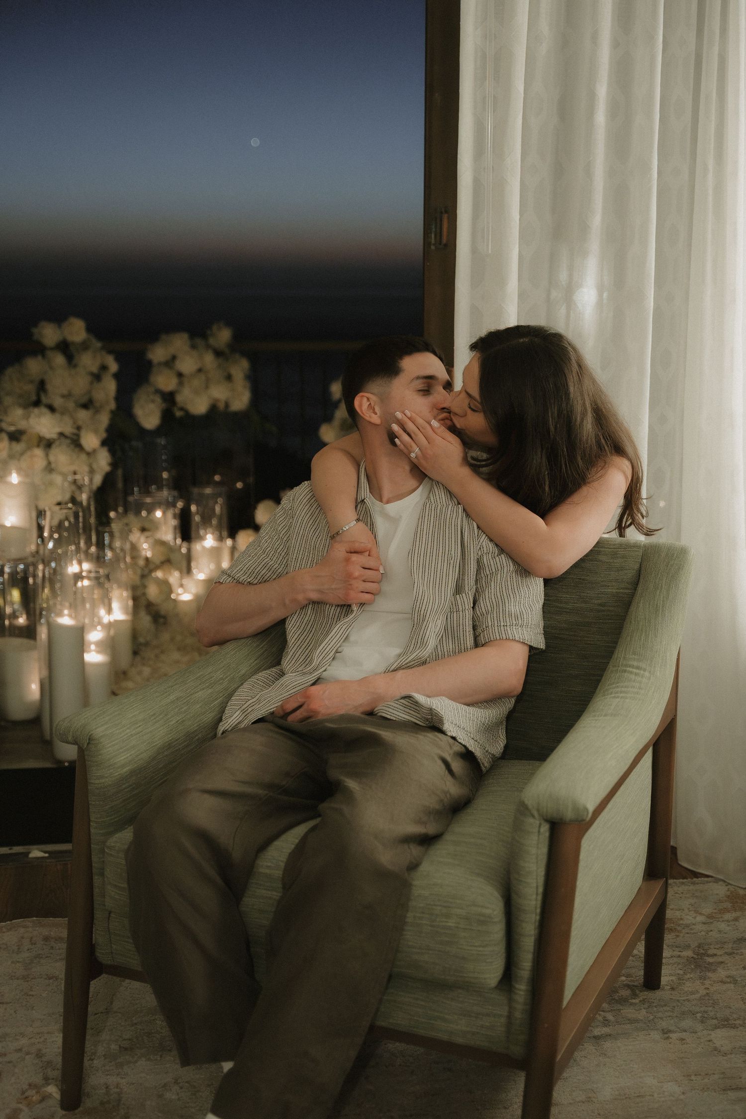 Romantic couple kissing on a green armchair surrounded by candles and white flowers at night.