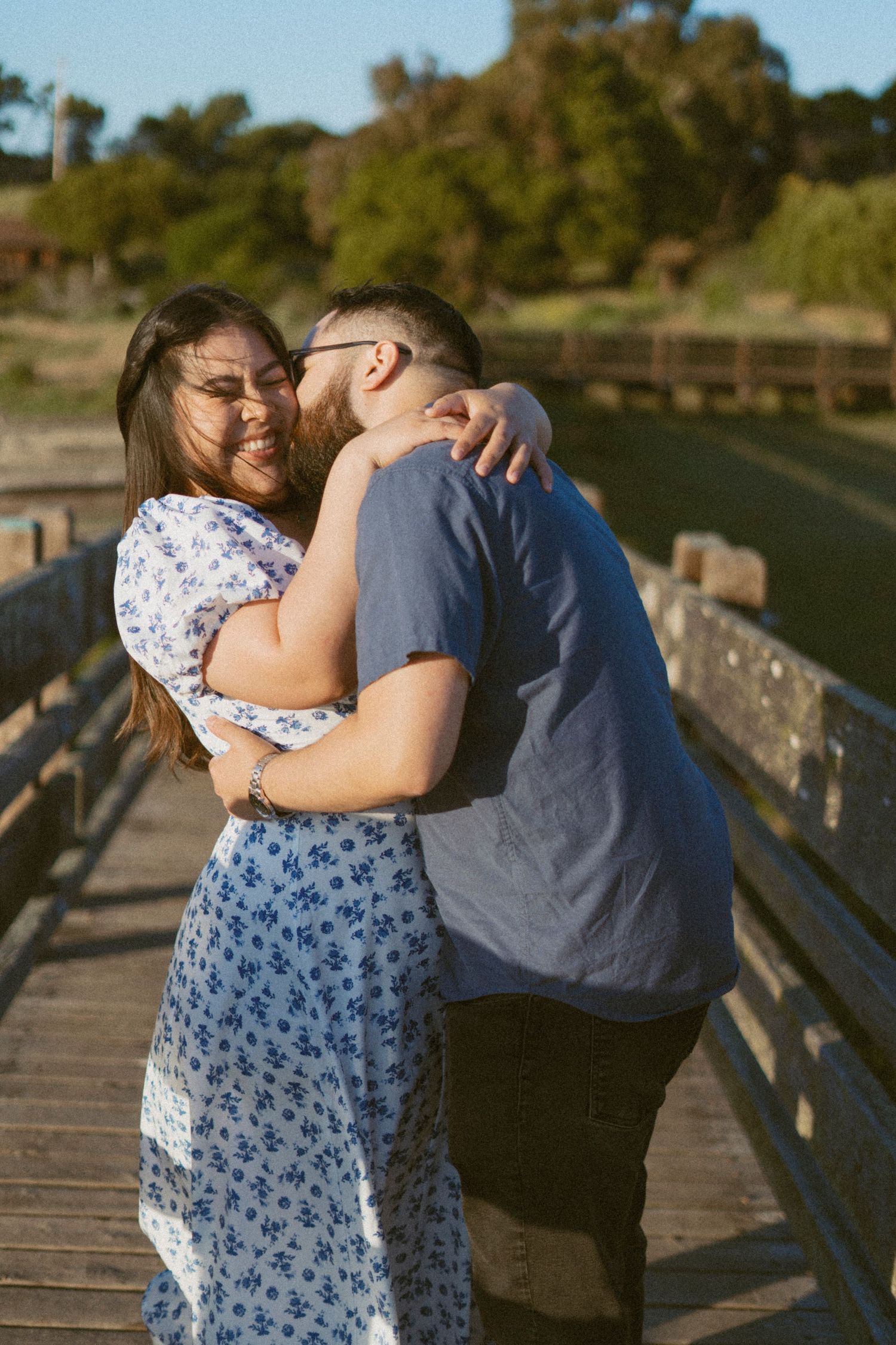 Happy couple embracing on a wooden bridge in warm golden sunlight with green trees and water in background in Fremont, CA.