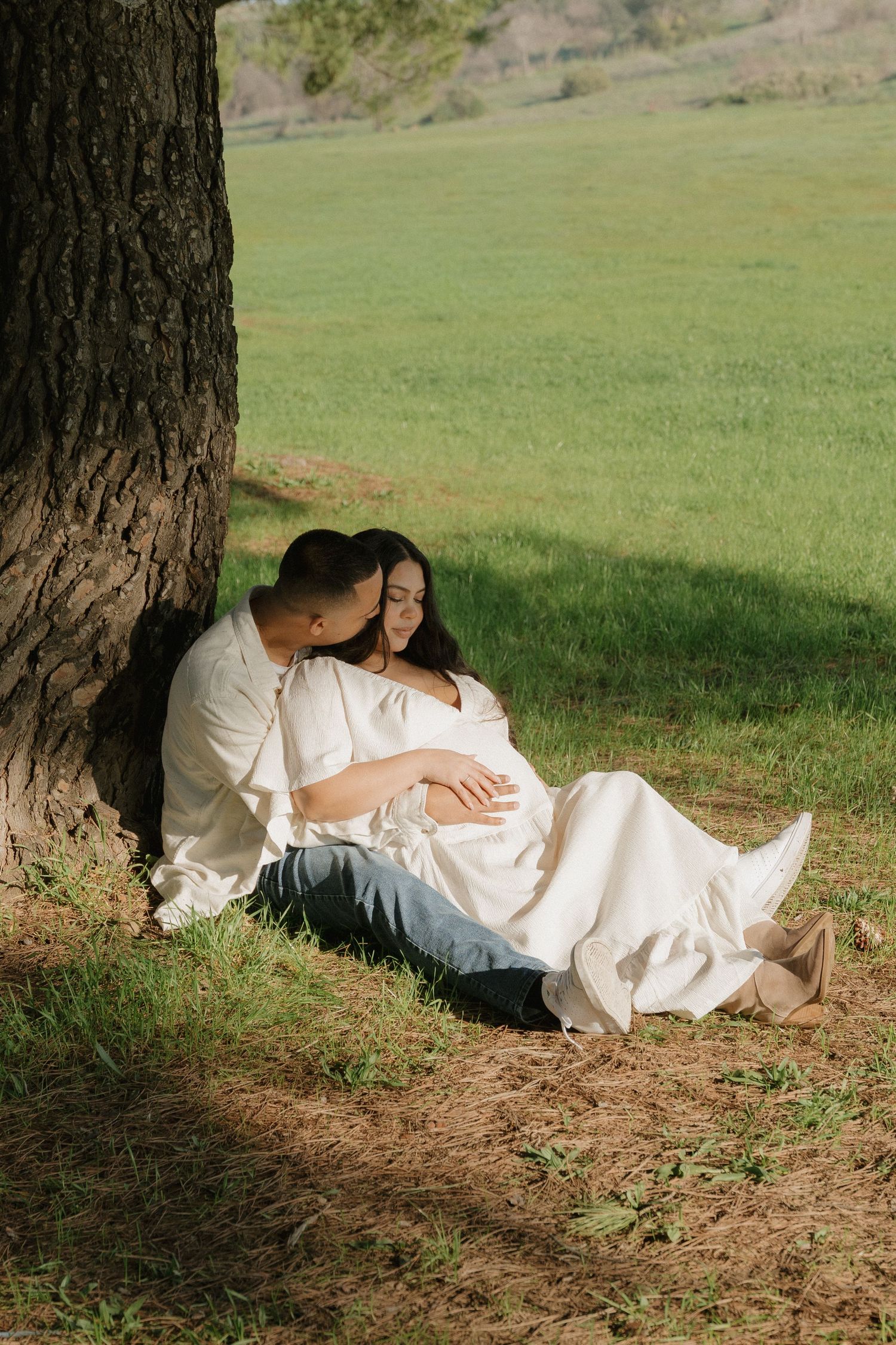 Pregnant couple sitting together against a large tree trunk in a sunny green field.
