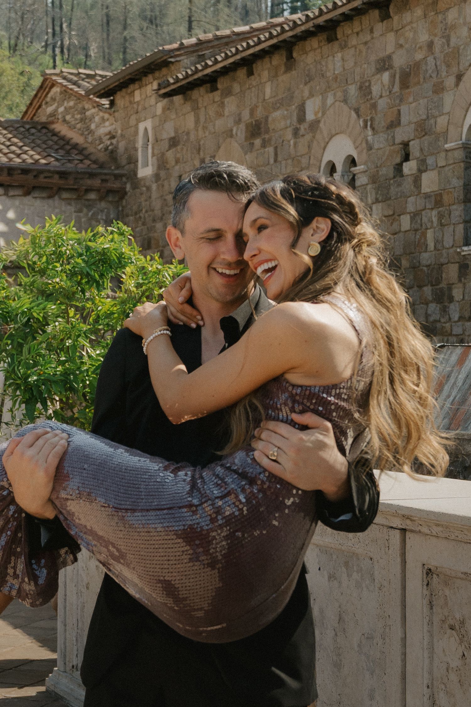 Happy couple laughing outdoors as man carries woman in sparkly sequin dress near stone building, at Napa.
