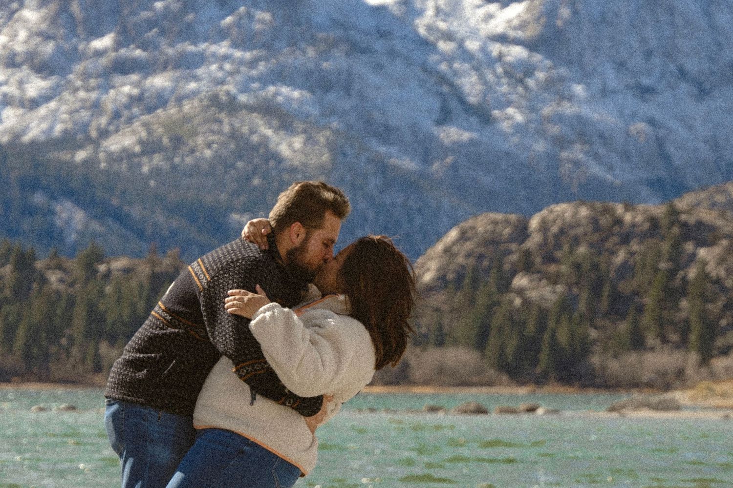 Couple sharing a romantic kiss by a turquoise mountain lake with snow-capped peaks in the background.