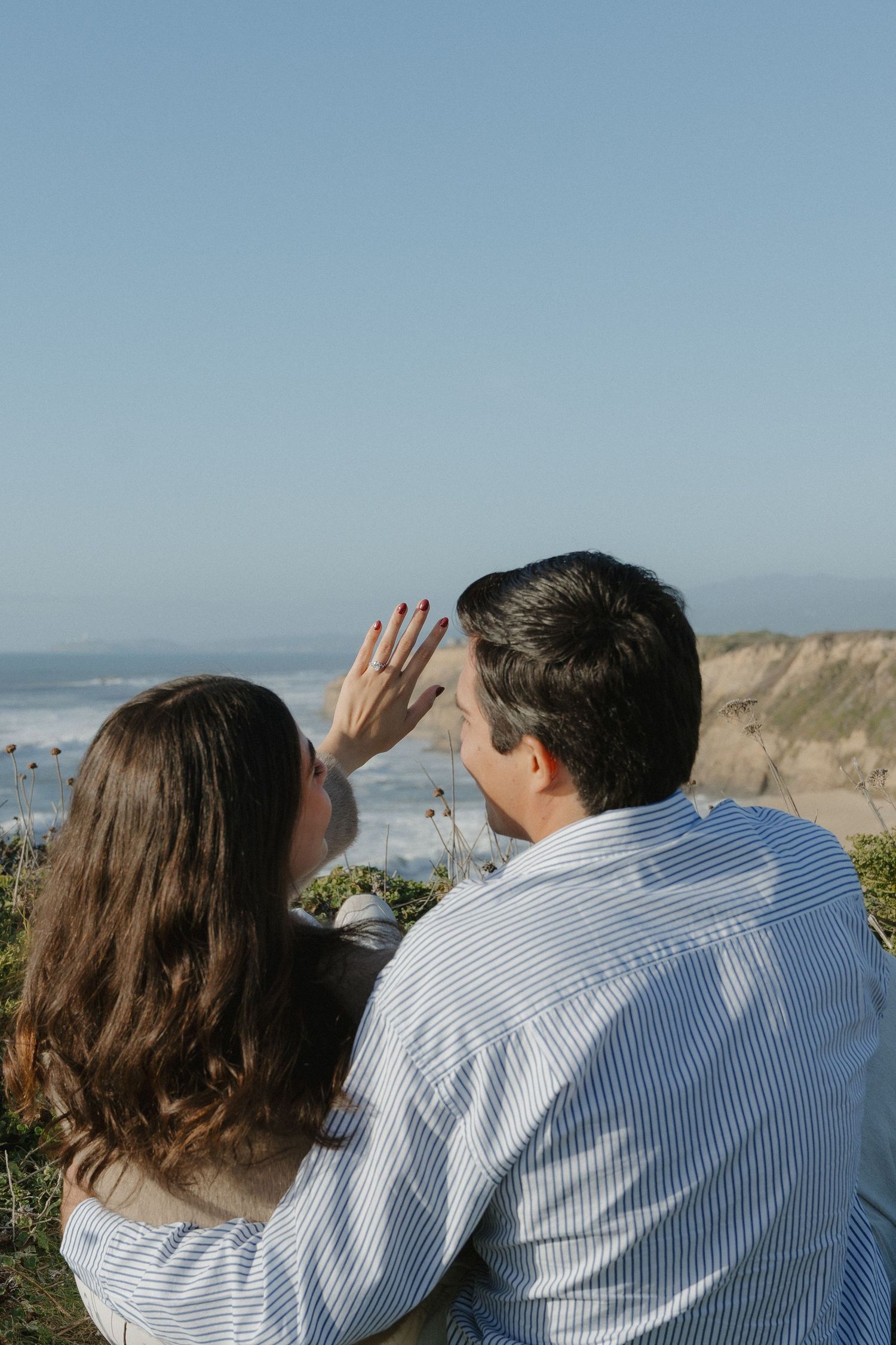 Engaged couple sitting together as woman shows off her ring with ocean cliffs in background at Half Moon Bay.