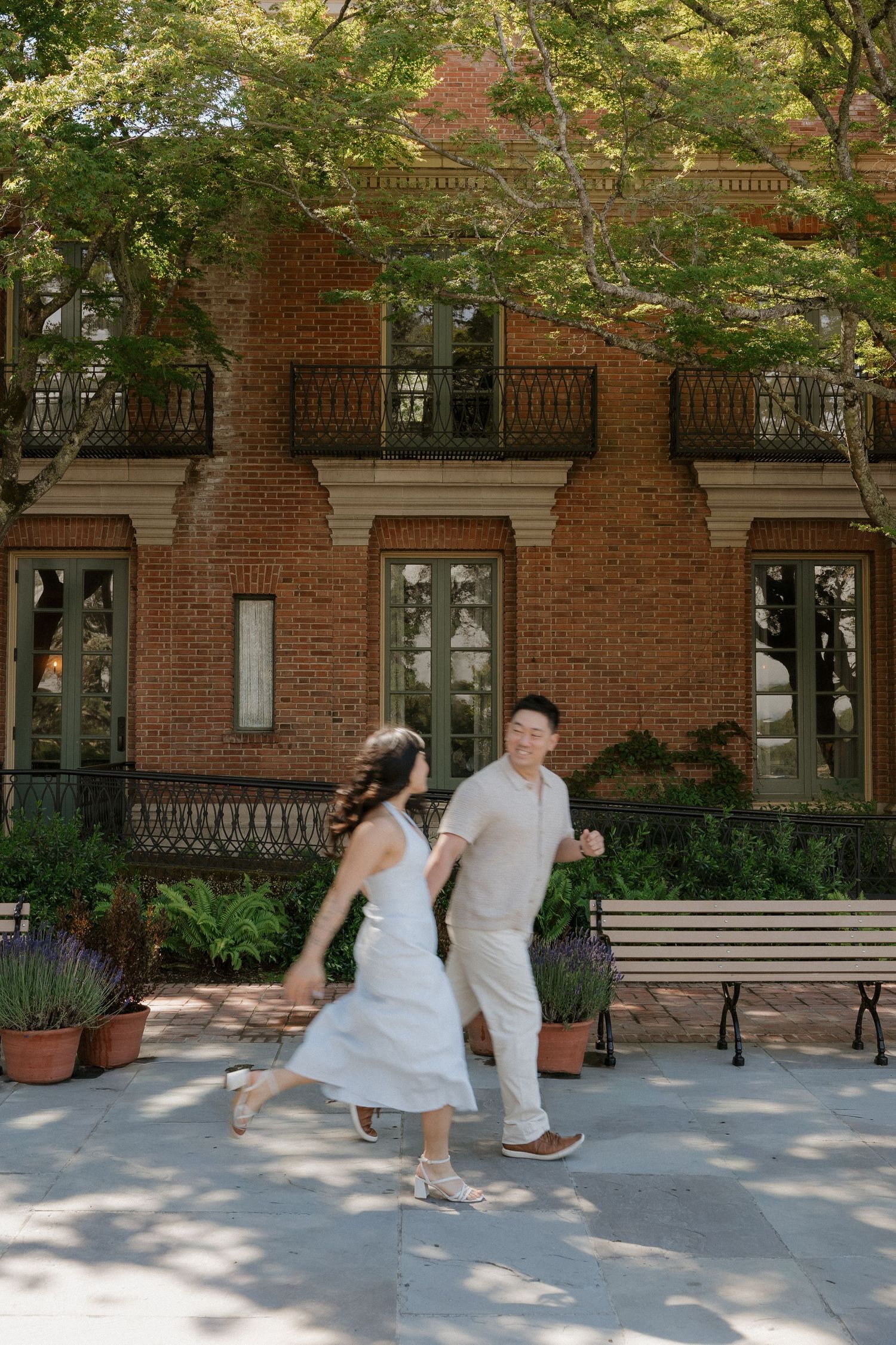 Couple in white outfits holding hands and running joyfully in front of a brick building with lush greenery.