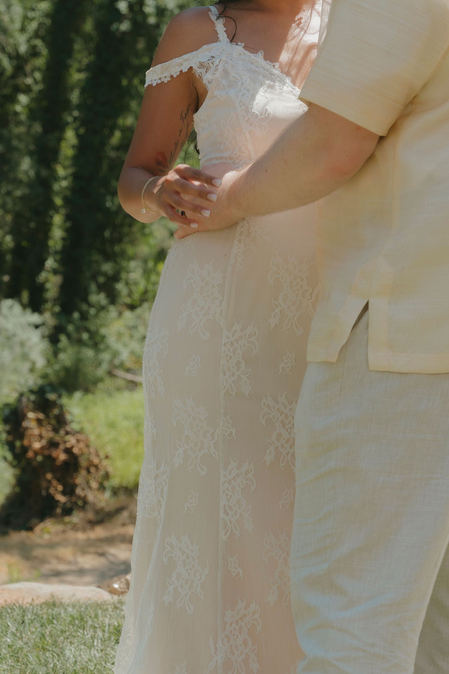 Close-up of bride in off-shoulder lace wedding gown being embraced by groom in cream suit outdoors.