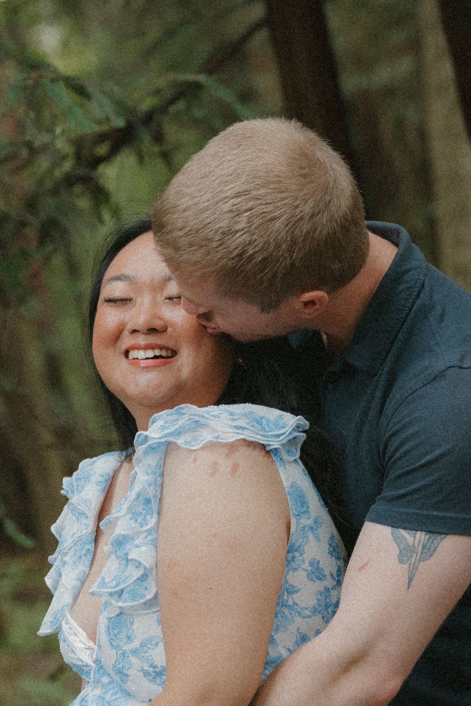 Joyful woman in blue floral dress laughing as her partner kisses her cheek in a lush green forest setting at Santa Cruz, CA.