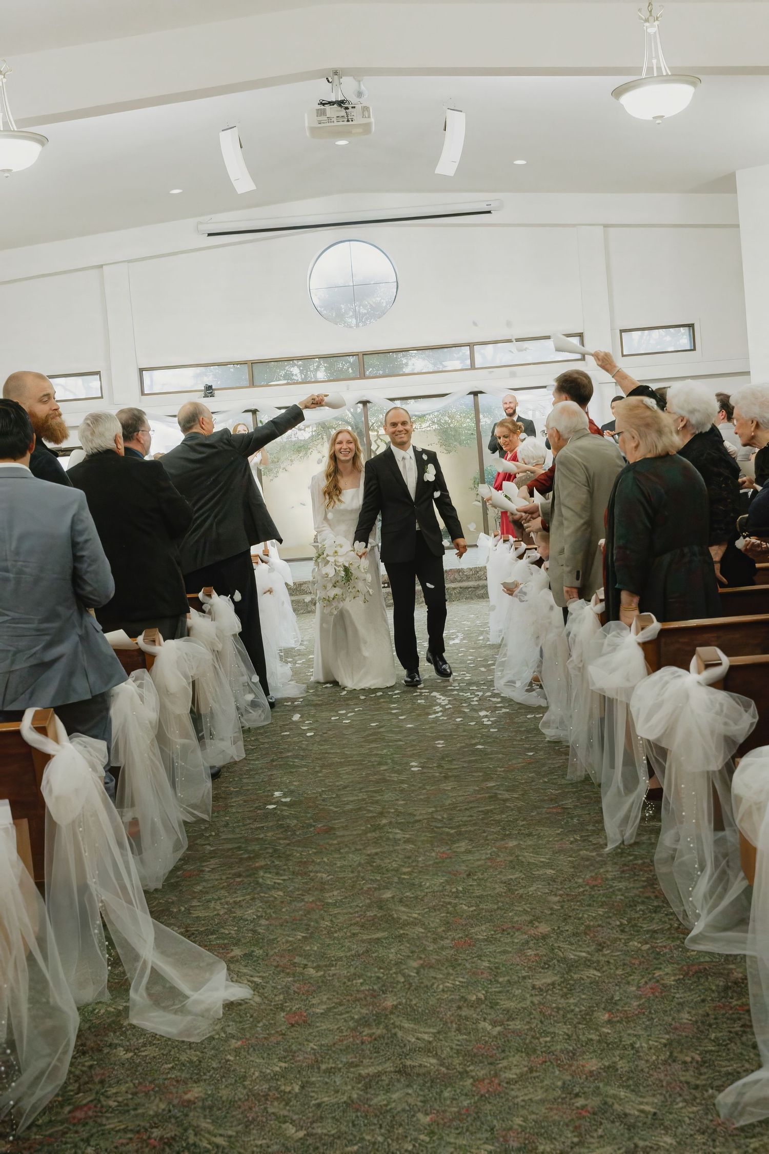 Newlyweds walking down the aisle of a white chapel as guests celebrate with confetti.